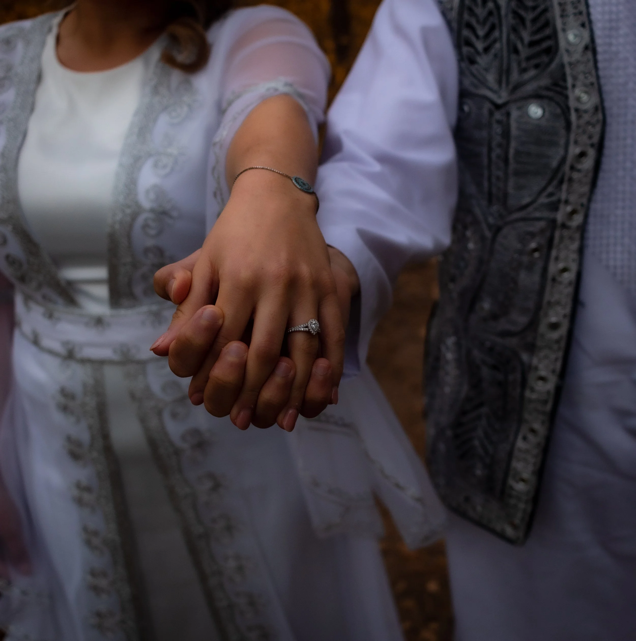 Close-up of a couple holding hands, showing wedding rings, with the woman wearing a white blouse with embroidery and a bracelet, and the man in traditional embroidered shirt.