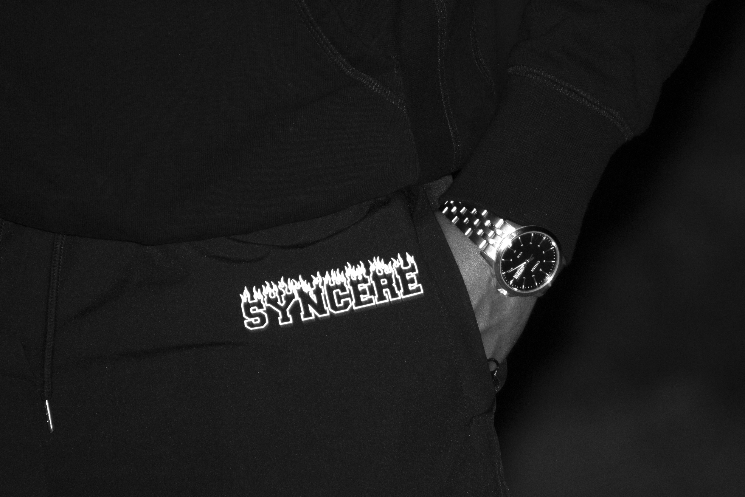 Close-up of a person wearing a black hoodie and black sweatpants with white graphic text, a silver wristwatch, and standing against a dark background.