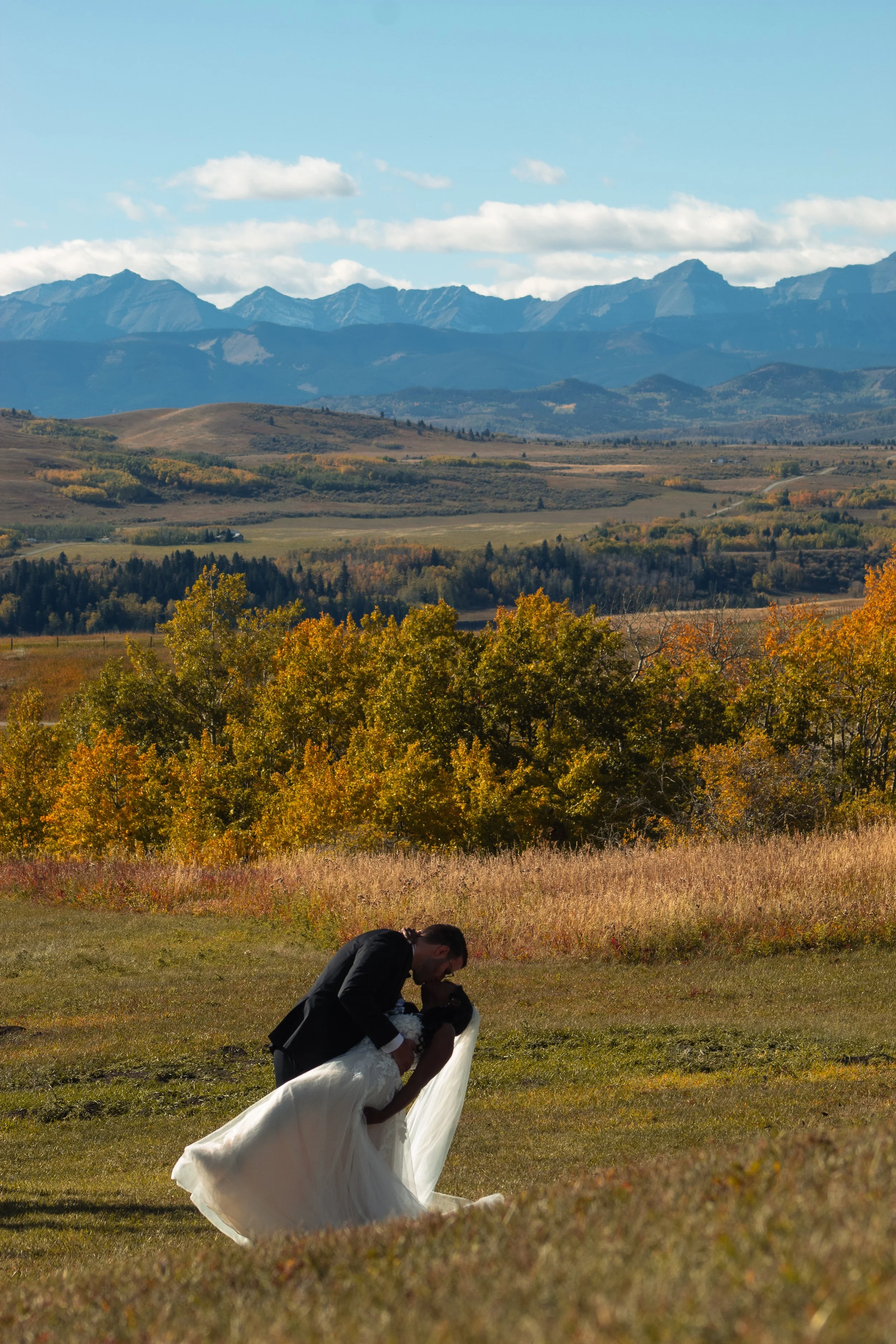 A person in formal attire, carrying a bride in a wedding gown, is kneeling on the grass in an outdoor setting with mountains, trees with autumn foliage, and a partly cloudy sky in the background.