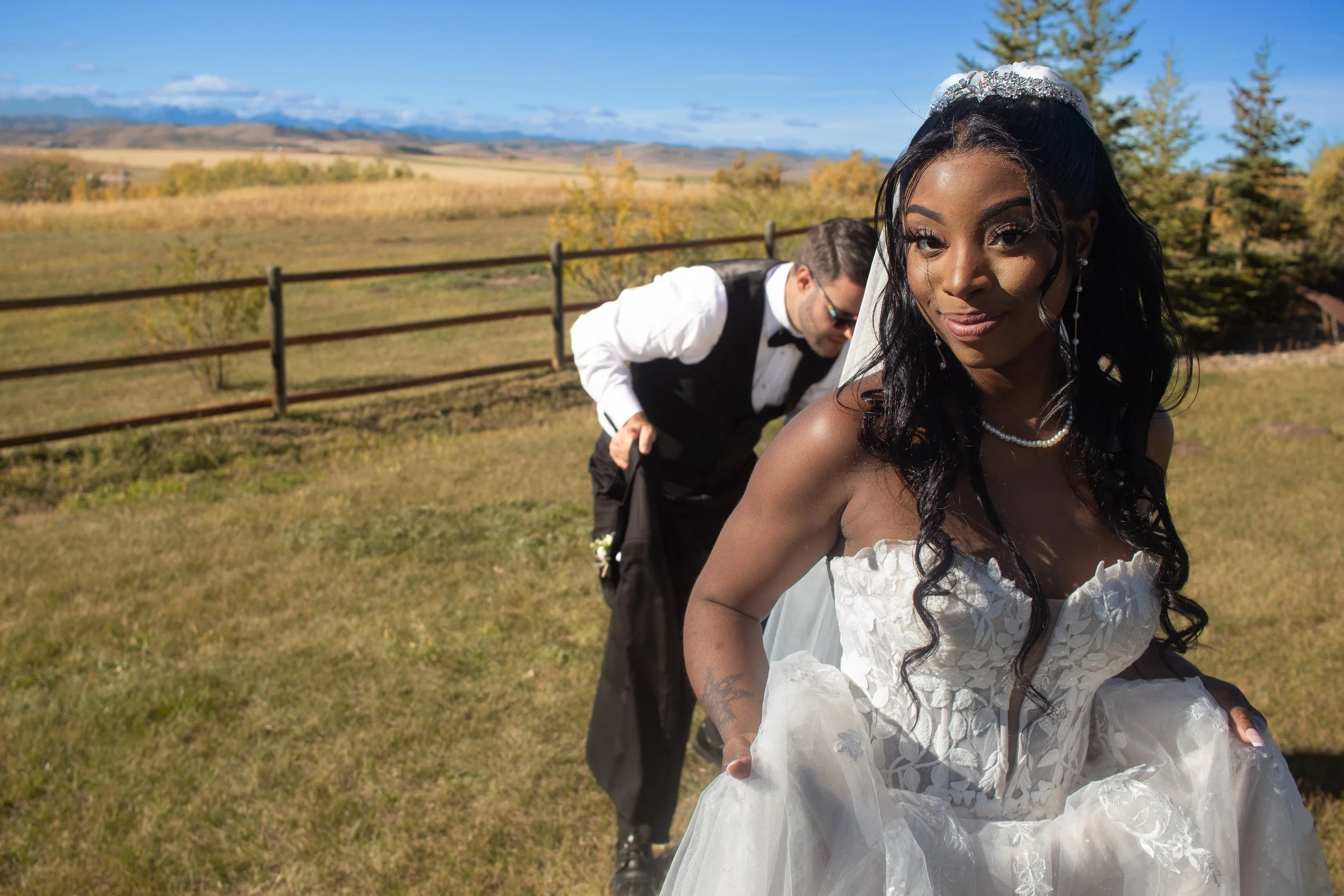 A bride and groom outdoors during the day, with the bride in a white wedding dress and the groom adjusting his suit behind her. The background shows a scenic view of open fields, trees, and mountains under a blue sky.