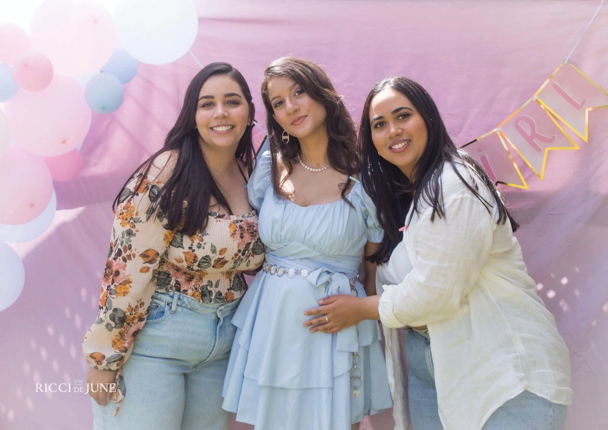 Three women smiling and posing together at a celebration with pink backdrop, balloons, and a banner.