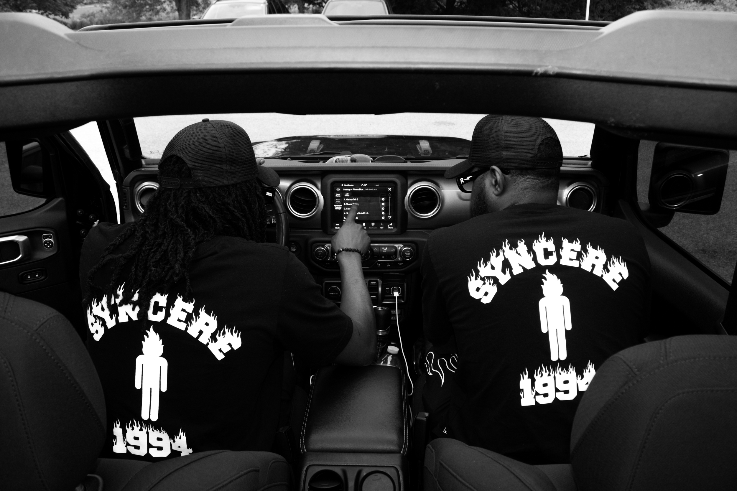 The image shows the interior of a car with two men sitting in front seats. Both men are wearing black t-shirts with graphic designs and the word 'SINCER' on the back. One man is pointing at a screen on the dashboard, which displays music options. The