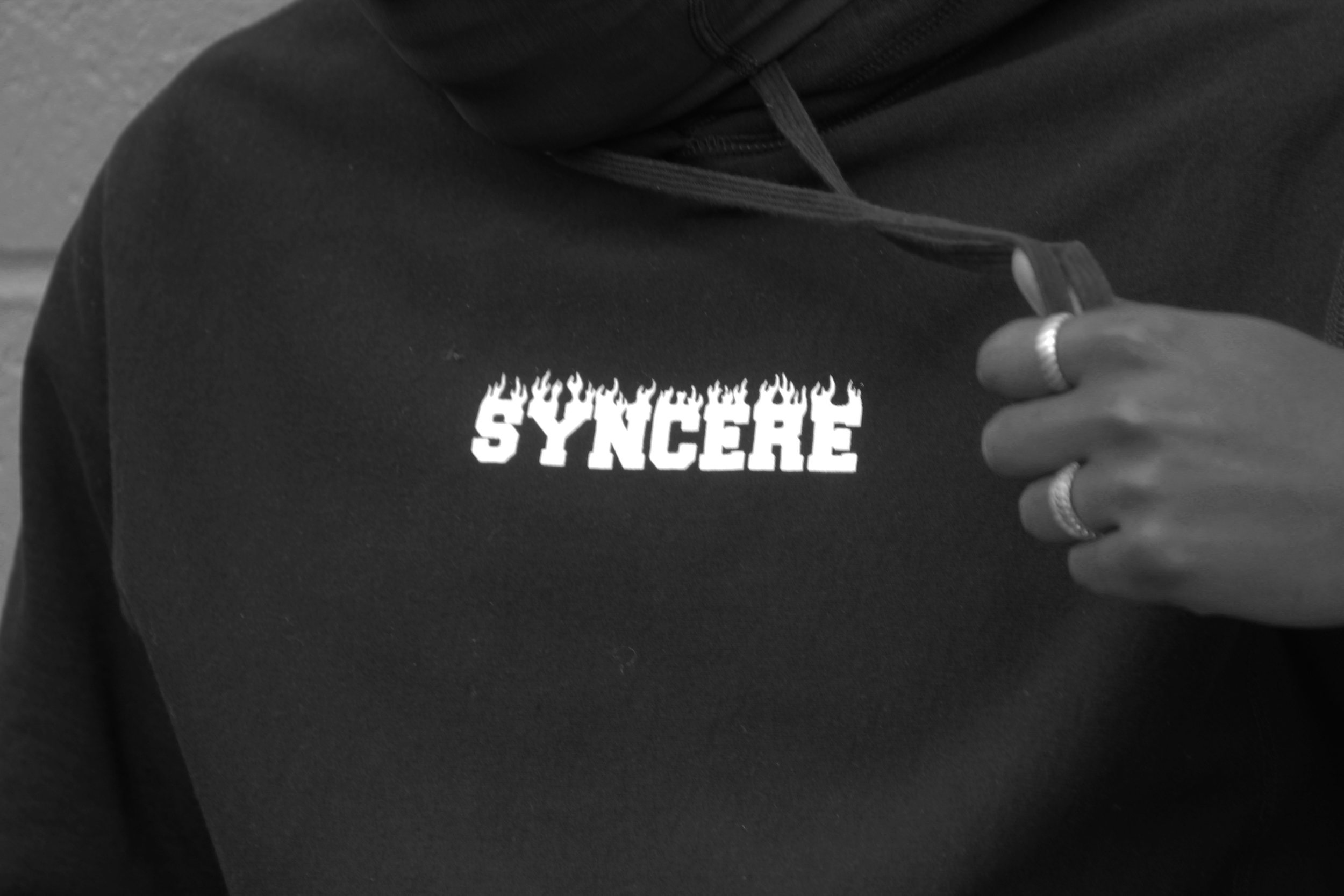 Close-up of a person wearing a black hoodie with the word 'SYNCRÉ' in fire-like letters on the chest. The person is holding the hoodie near the chest with fingers, wearing rings, and a chain necklace is visible. The background is blurred.