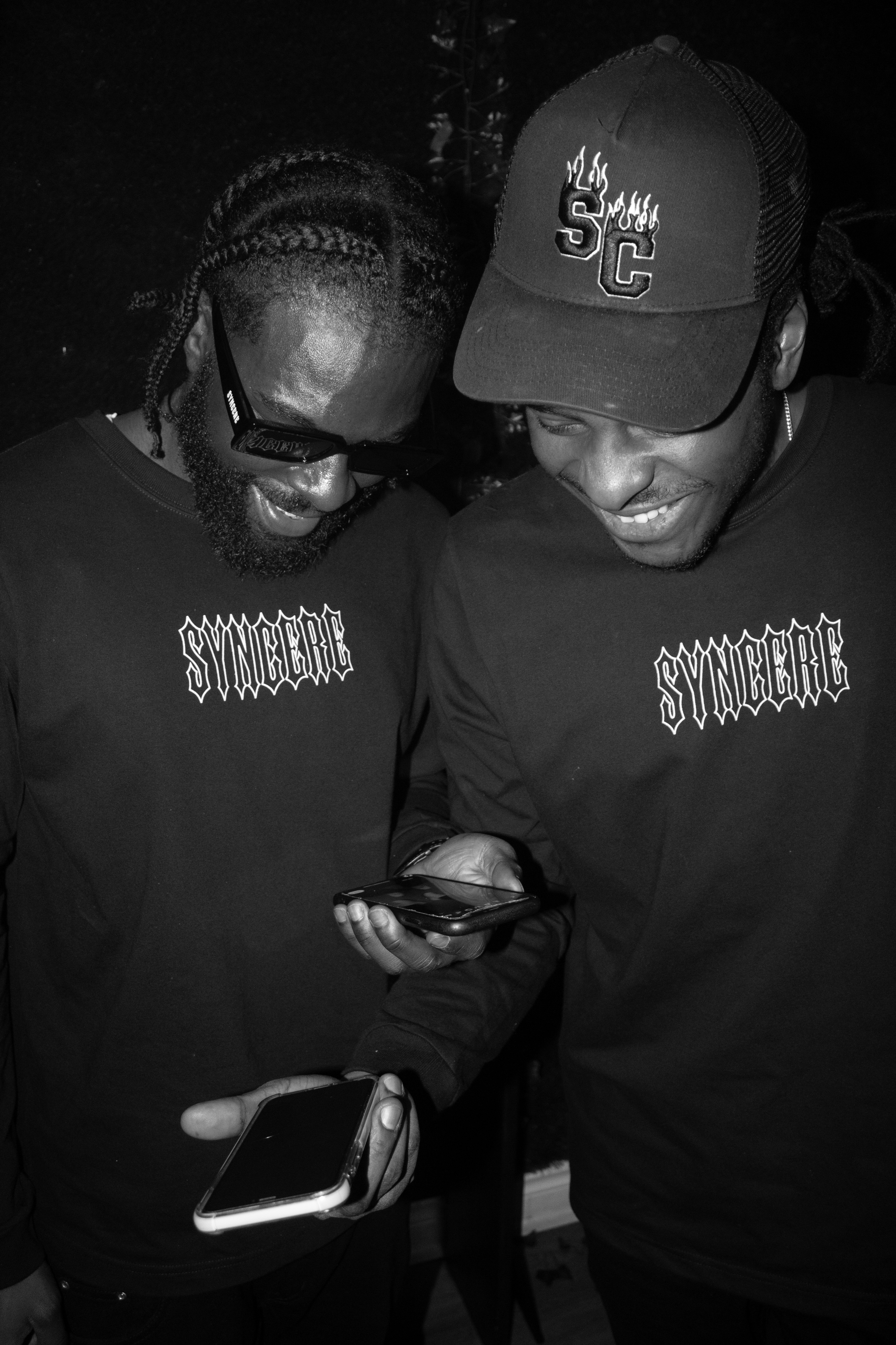 Two men looking at a smartphone together, both wearing dark shirts with the word "SYNCHRE" printed on them. One man is wearing a baseball cap with "SC" and flames embroidered on the front. The other man is wearing glasses and a watch.