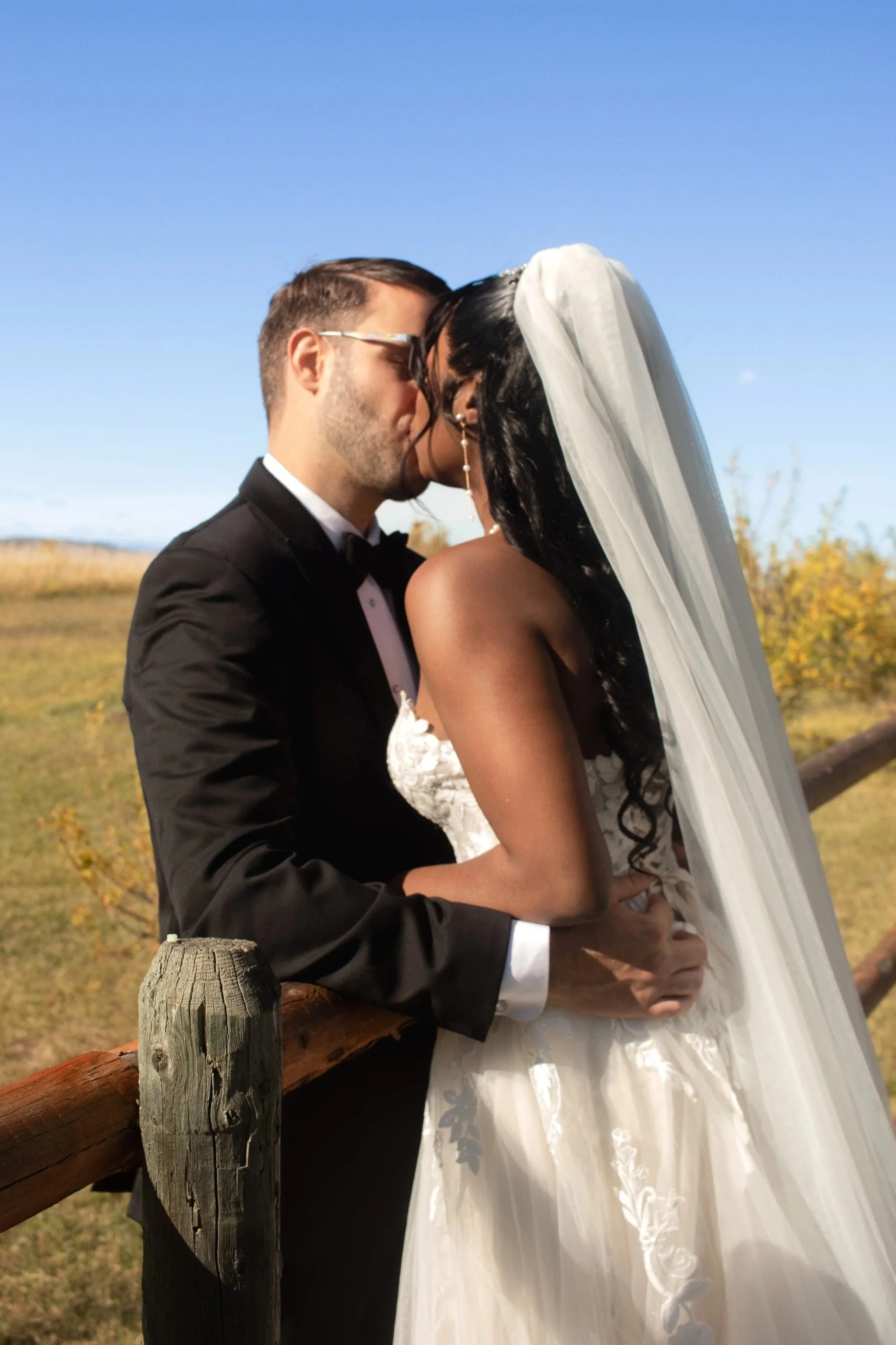 A couple is sharing a kiss outdoors during a wedding ceremony. The man is wearing a tuxedo and the woman is dressed in a wedding gown with a veil. They are standing next to a wooden fence with a scenic background of grass, trees, and a clear sky.
