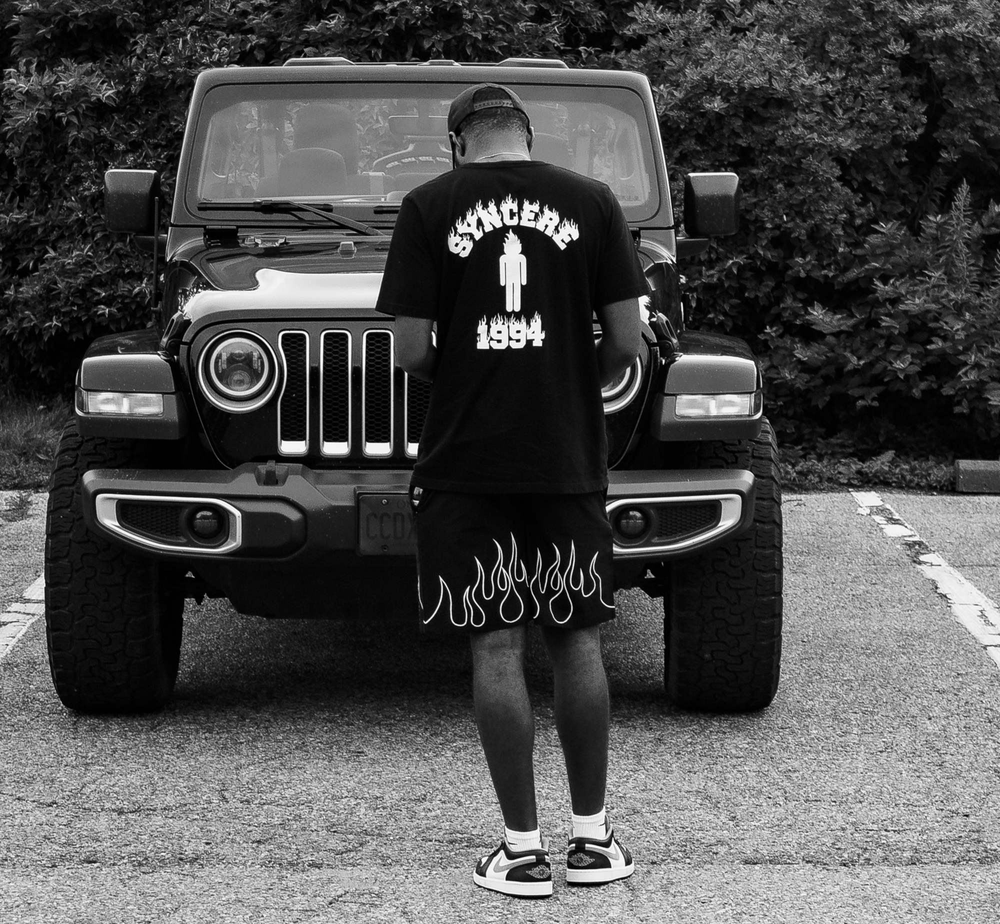 Black and white photo of a person standing in front of a Jeep, with their back turned towards the camera. The person is wearing a t-shirt with the word 'Sánchez' and the year '1994' on the back, along with a graphic of a burning file. They are also w