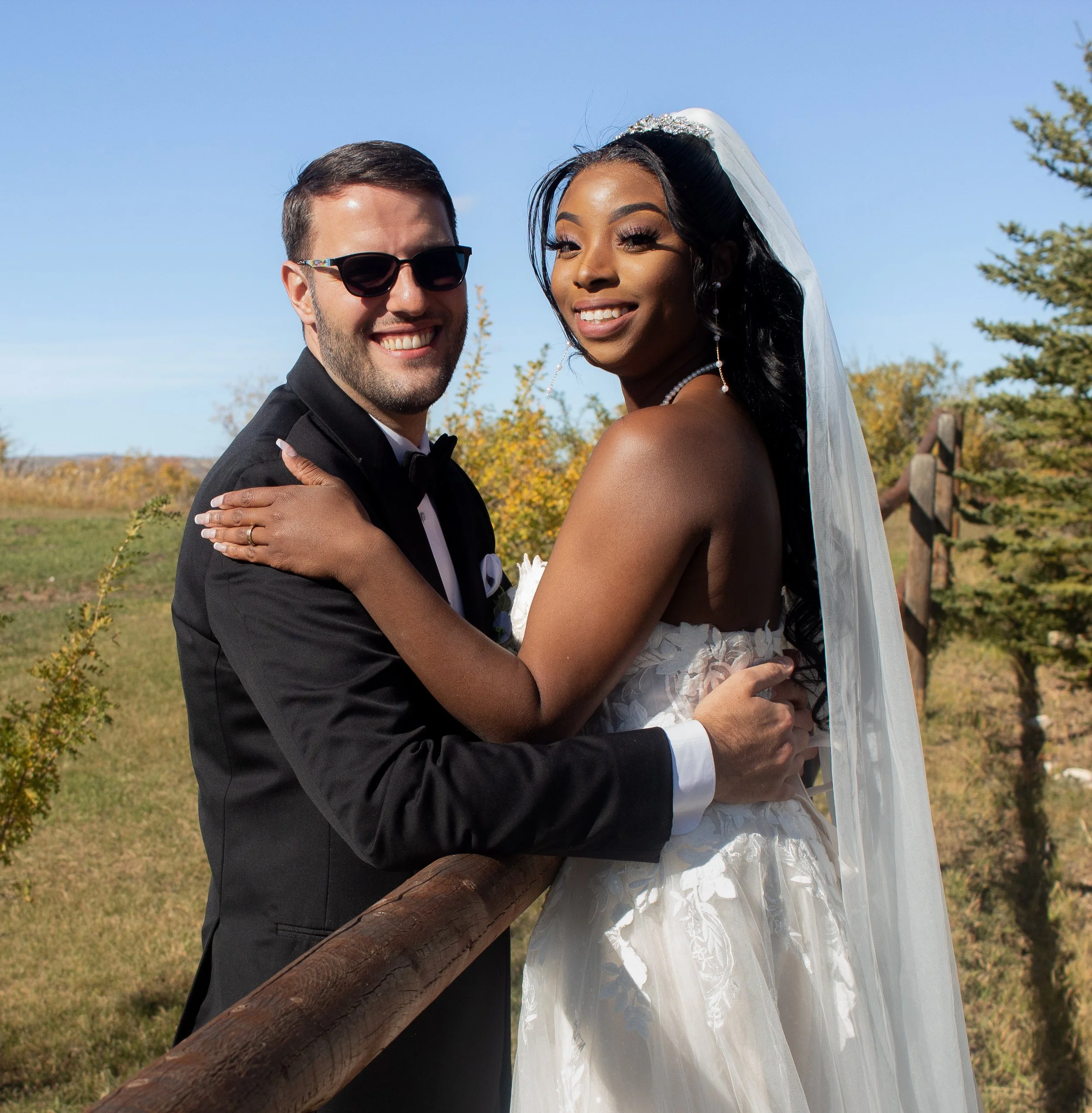 A happy couple in wedding attire, standing outdoors, with the groom in a tuxedo and the bride in a white wedding dress and veil, smiling at the camera.