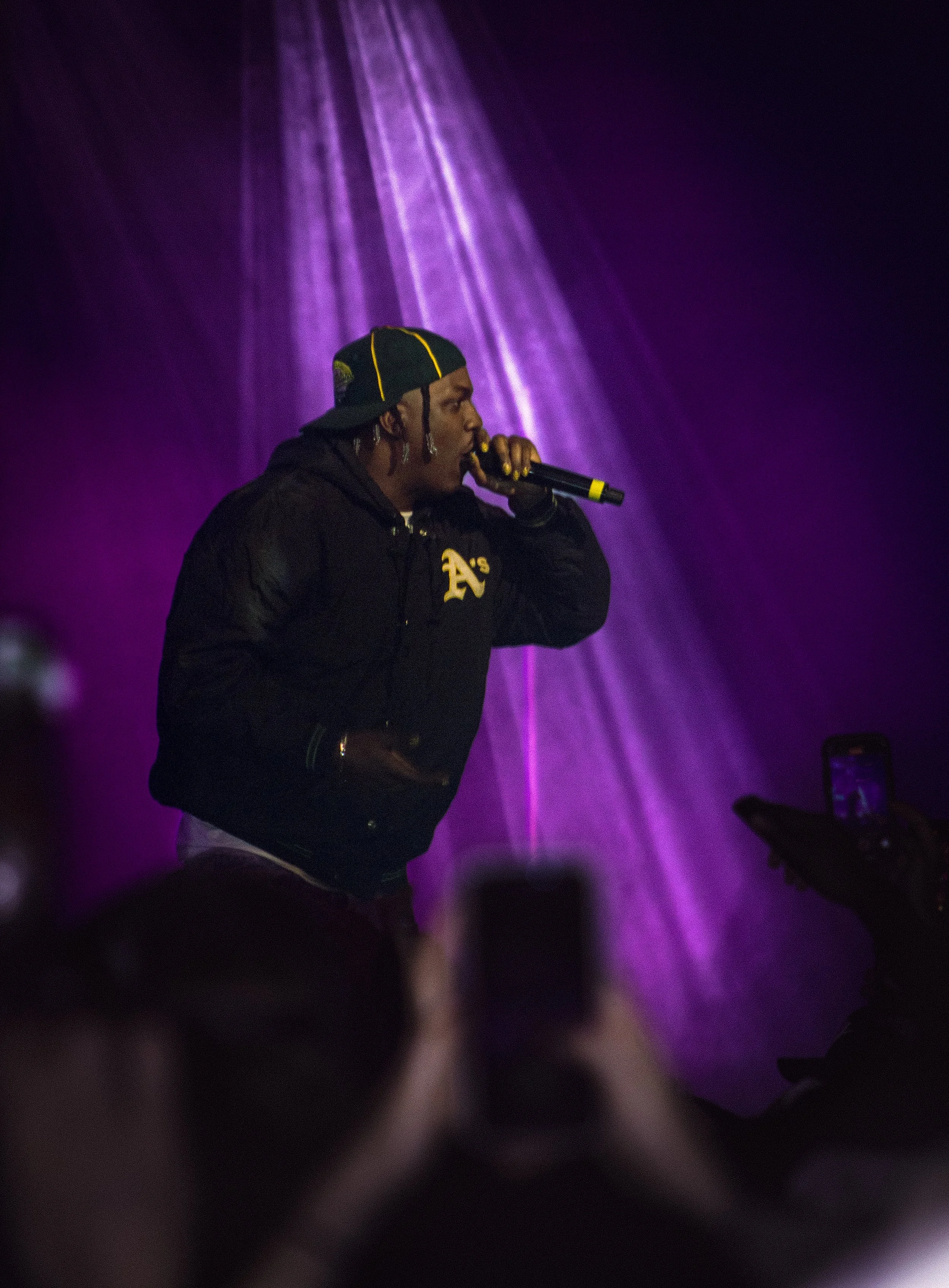 A person with braided hair, wearing a black cap backward and a black jacket, holding a microphone and performing on stage with purple lighting.