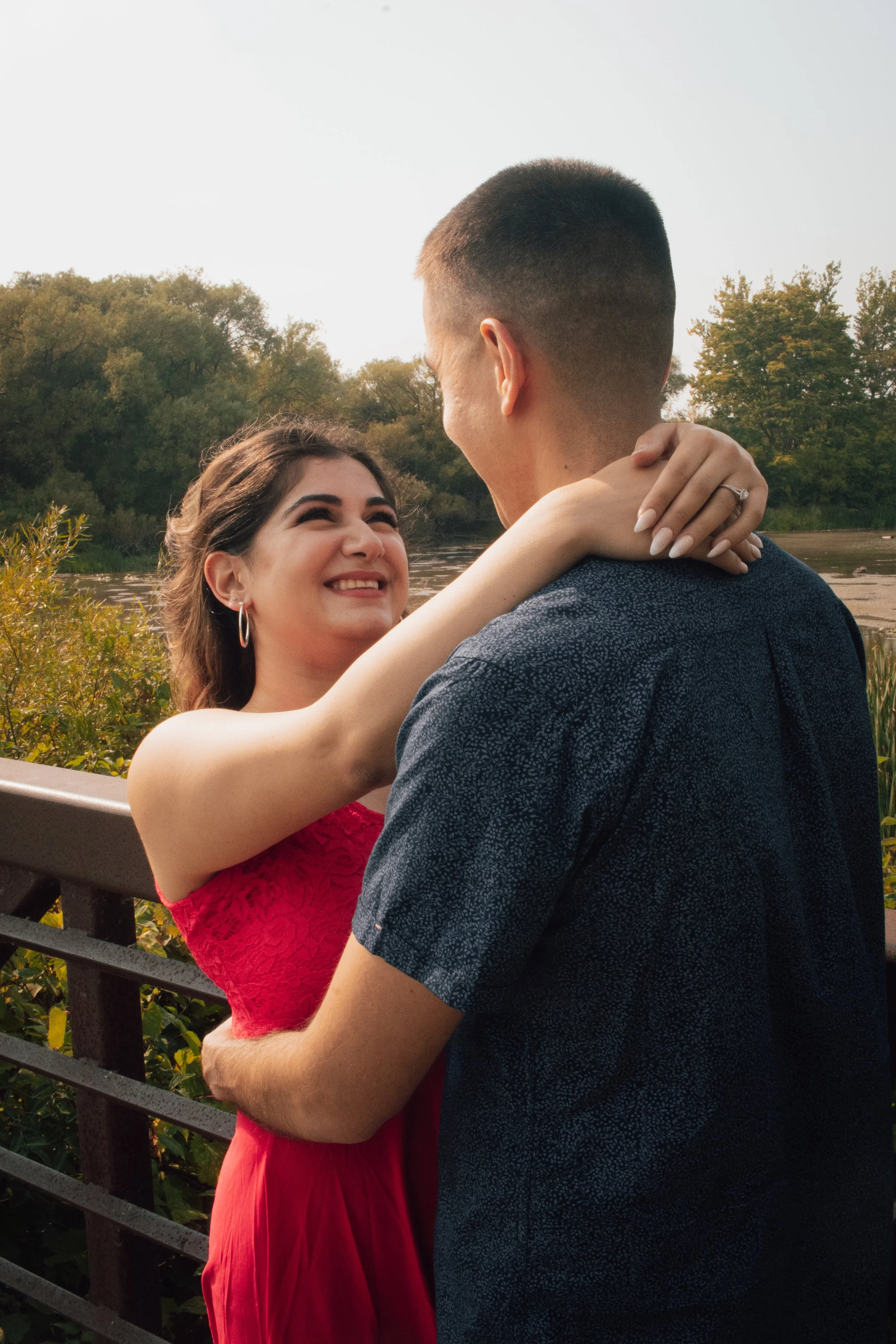 Couple embracing outdoors near a river with trees in the background, woman in a red dress smiling at man, who is wearing a blue patterned shirt.