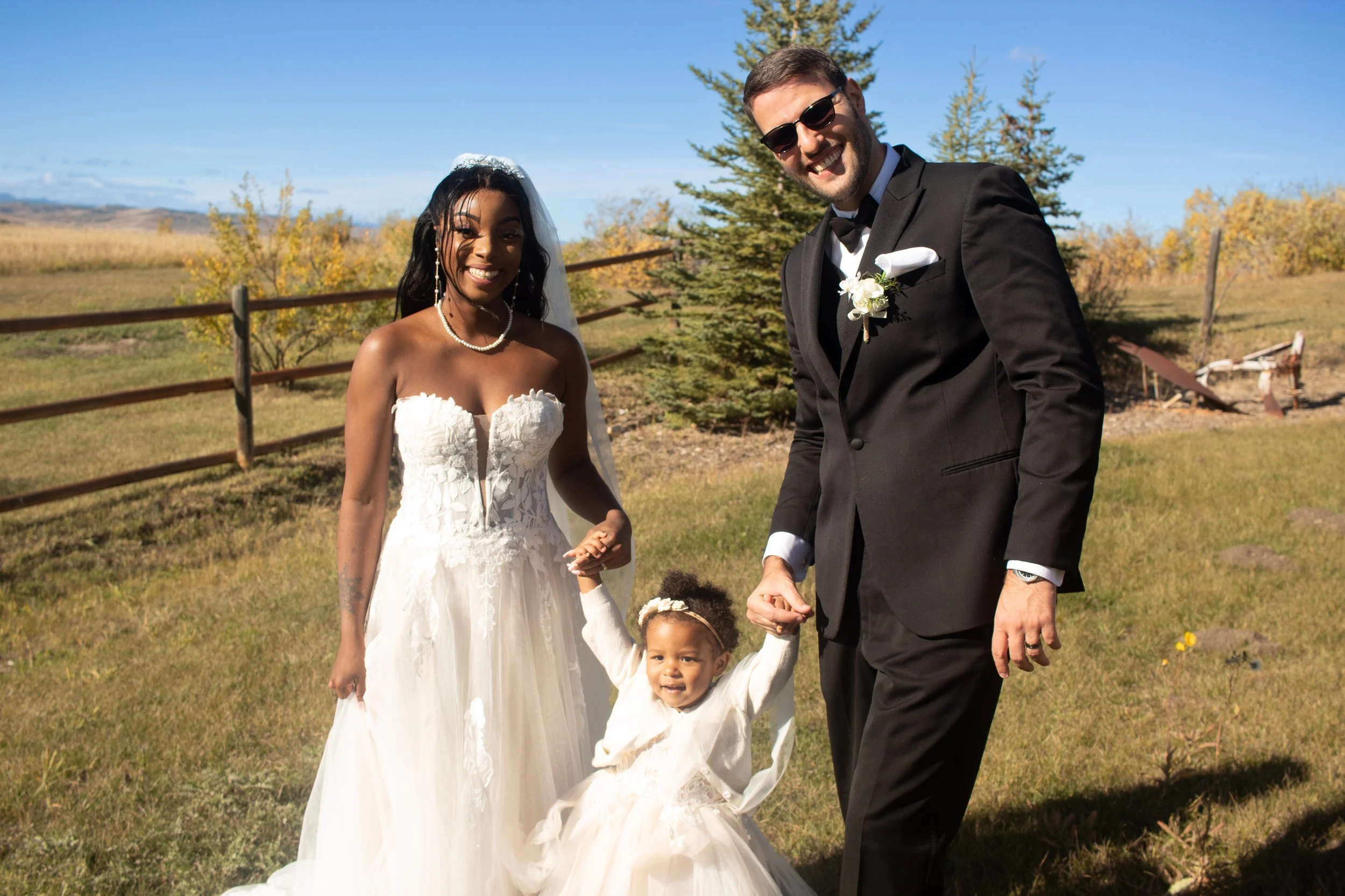 A bride and groom with two young children outside on a sunny day. The bride is wearing a white wedding dress and veil, and the groom in a black suit with sunglasses. The children are in white dresses, holding hands with the adults. There are trees an