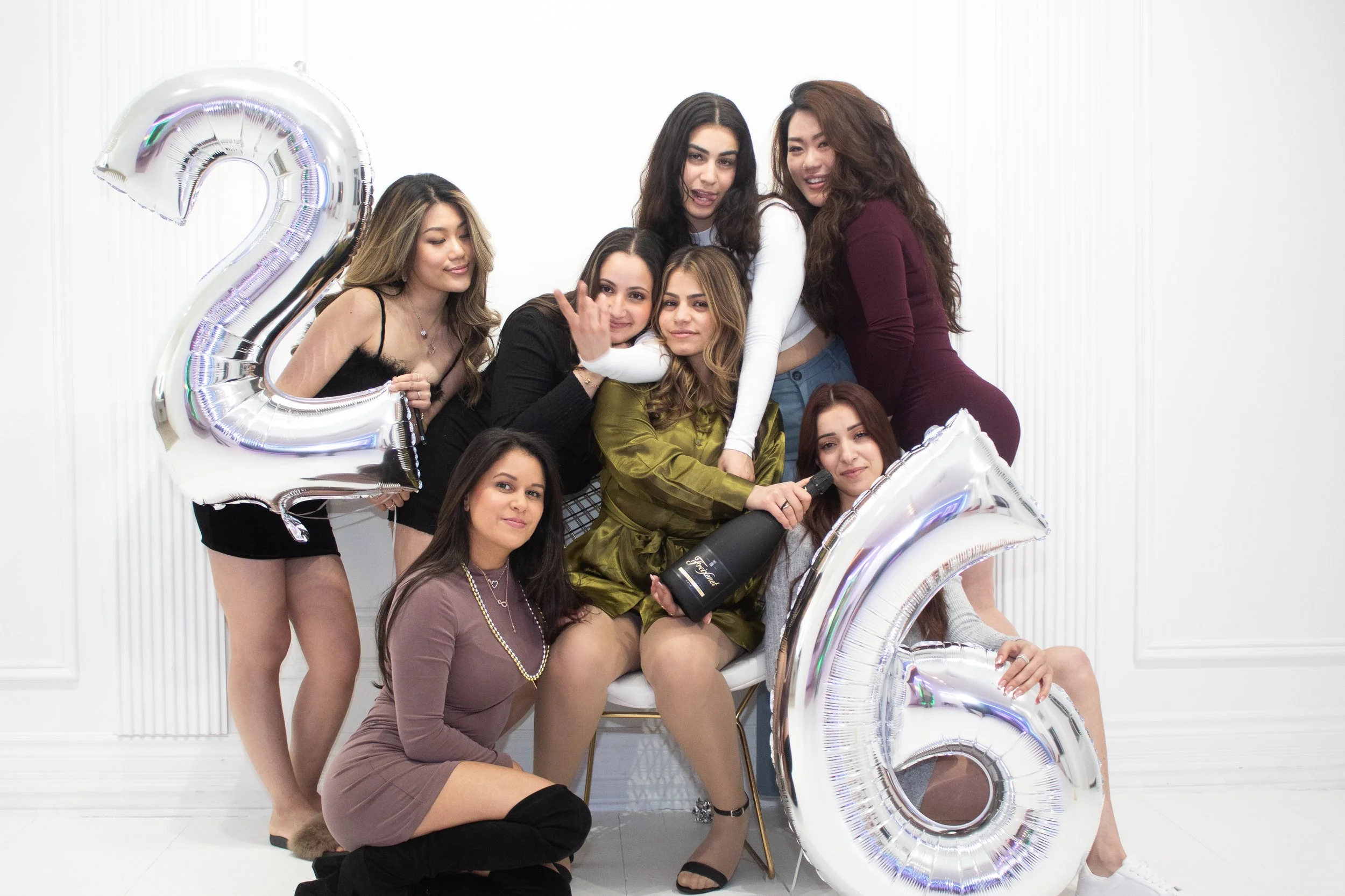 Group of women celebrating with balloons and champagne, holding a large silver number 2 balloon, in a white room.