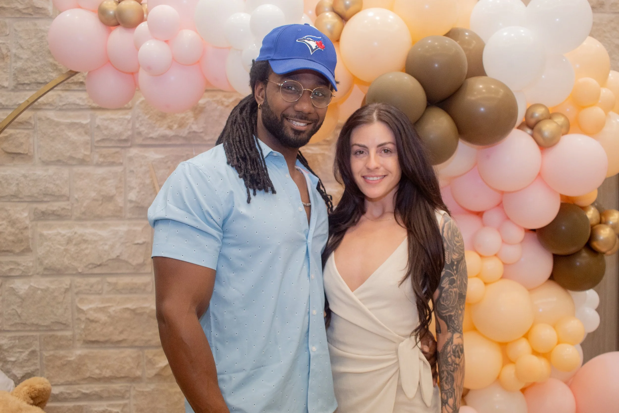 A man and woman smiling in front of a balloon backdrop with various pink, white, gold, and brown balloons, and a teddy bear to the left.