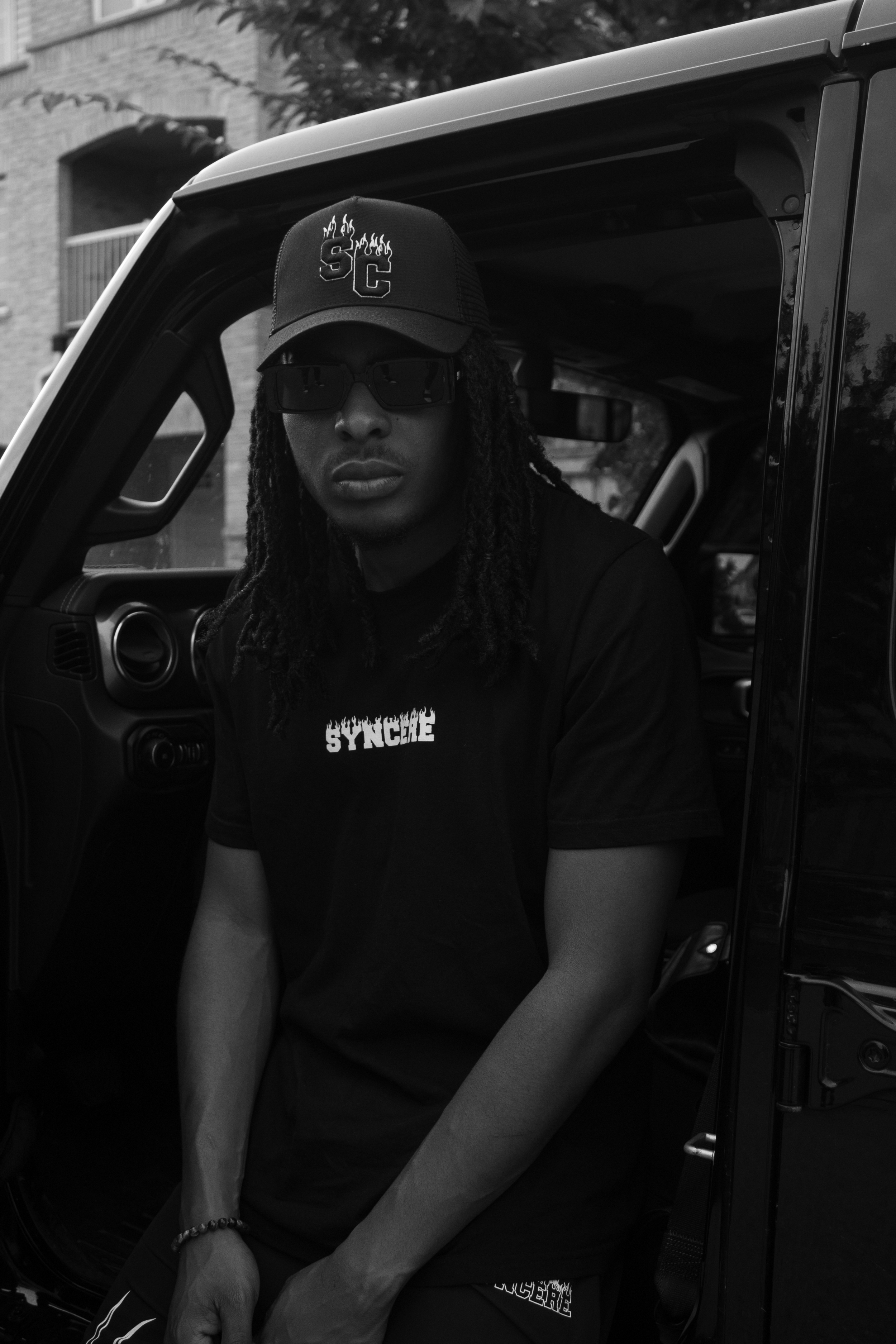 Young man with dreadlocks wearing sunglasses, a cap with flaming letters, and a black T-shirt with white text sitting in a car.