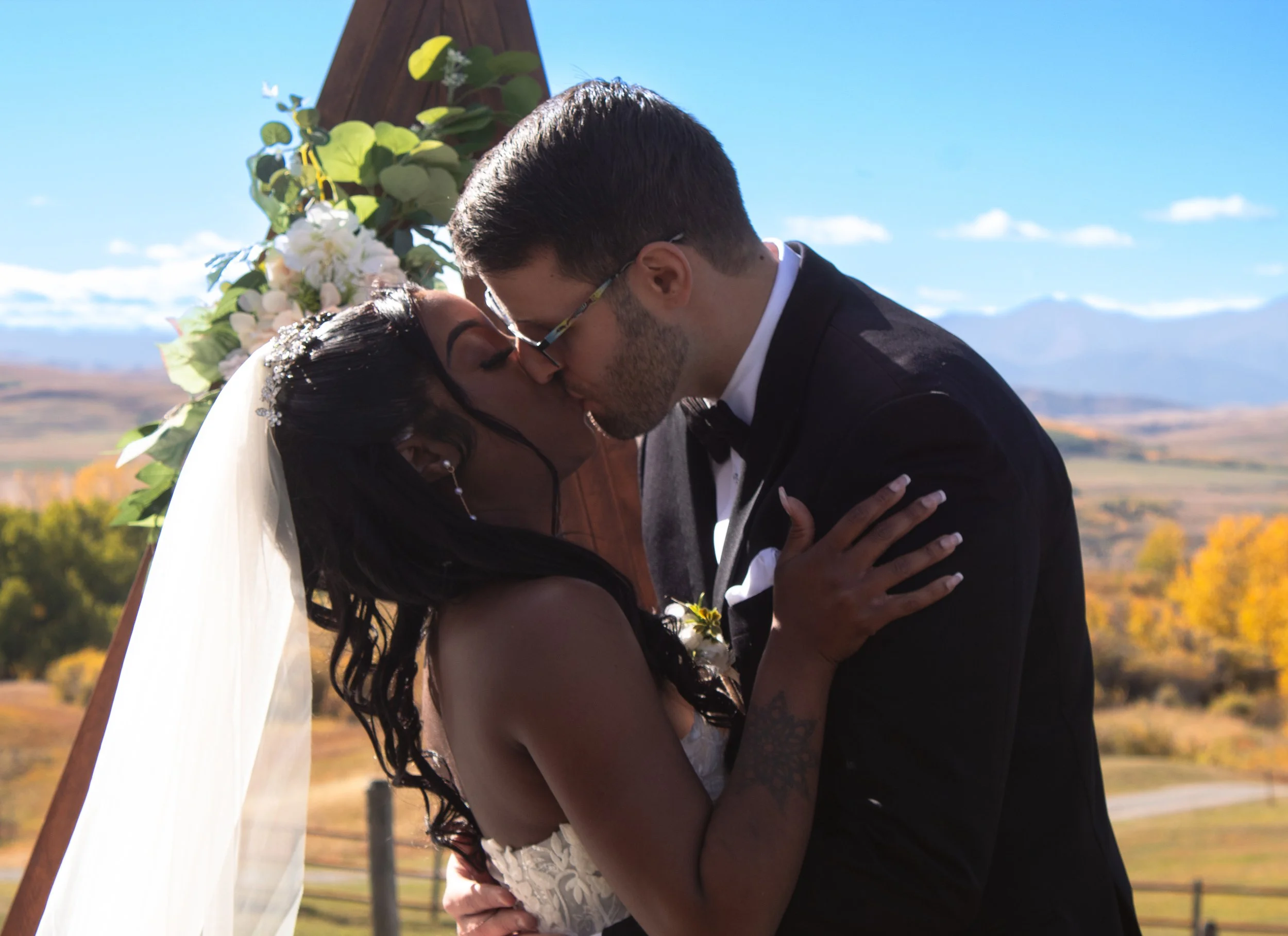 A newly married couple sharing a kiss outdoors under a wedding arch decorated with flowers and green leaves, with a scenic landscape of mountains and colorful trees in the background.