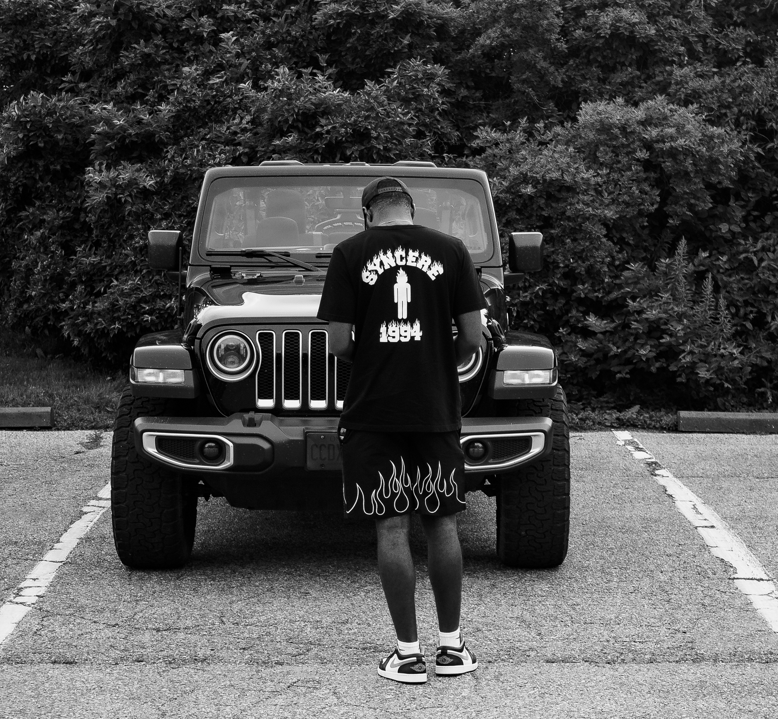 A man in a black T-shirt with flaming design and the text "Bulancer 1994" is standing in front of a Jeep Wrangler, looking down, with a background of dense foliage.