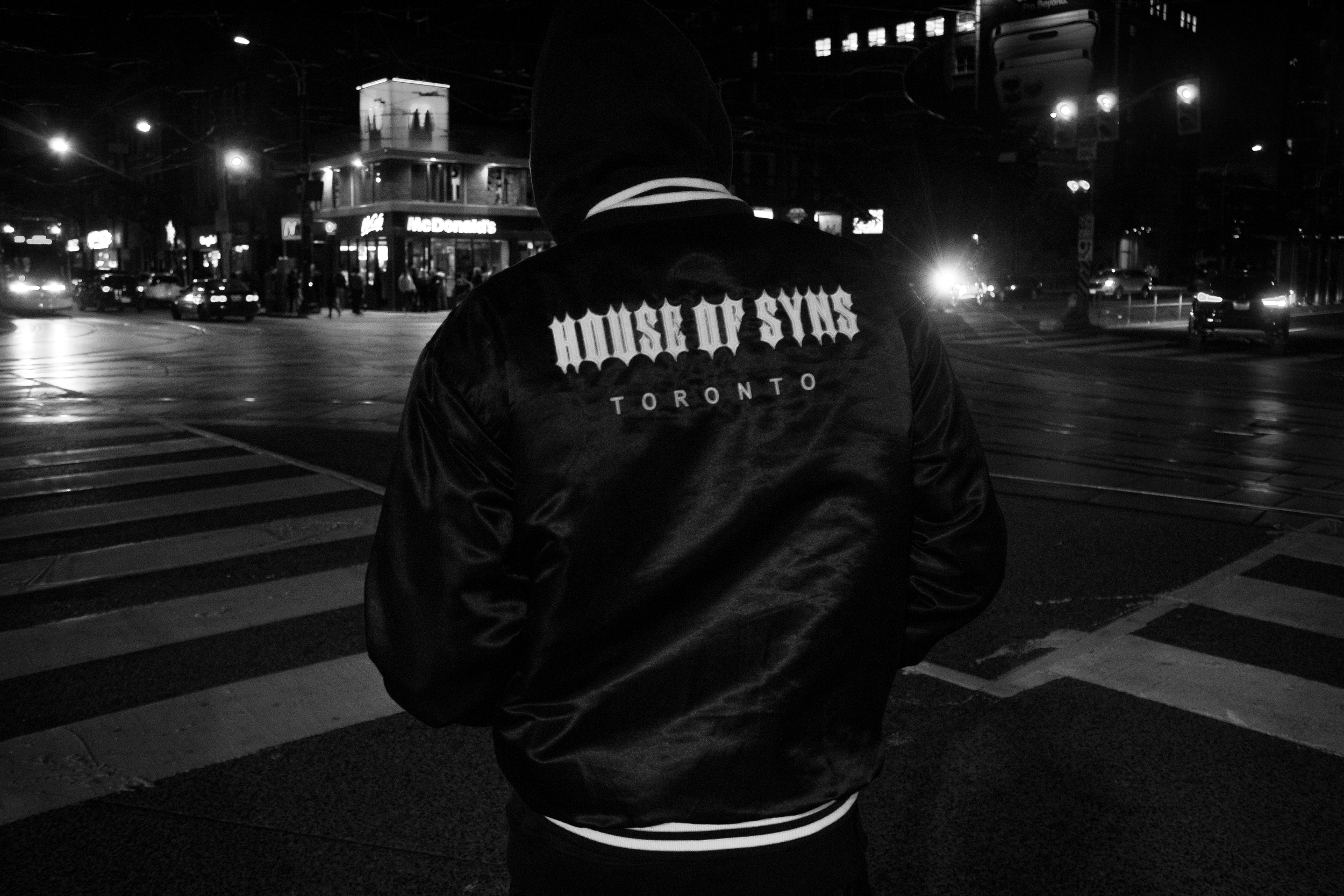 A person wearing a black jacket with 'HOUSE OF SYNS TORONTO' written on the back, standing at a crosswalk at night in an urban area with cars and a McDonald's in the background.