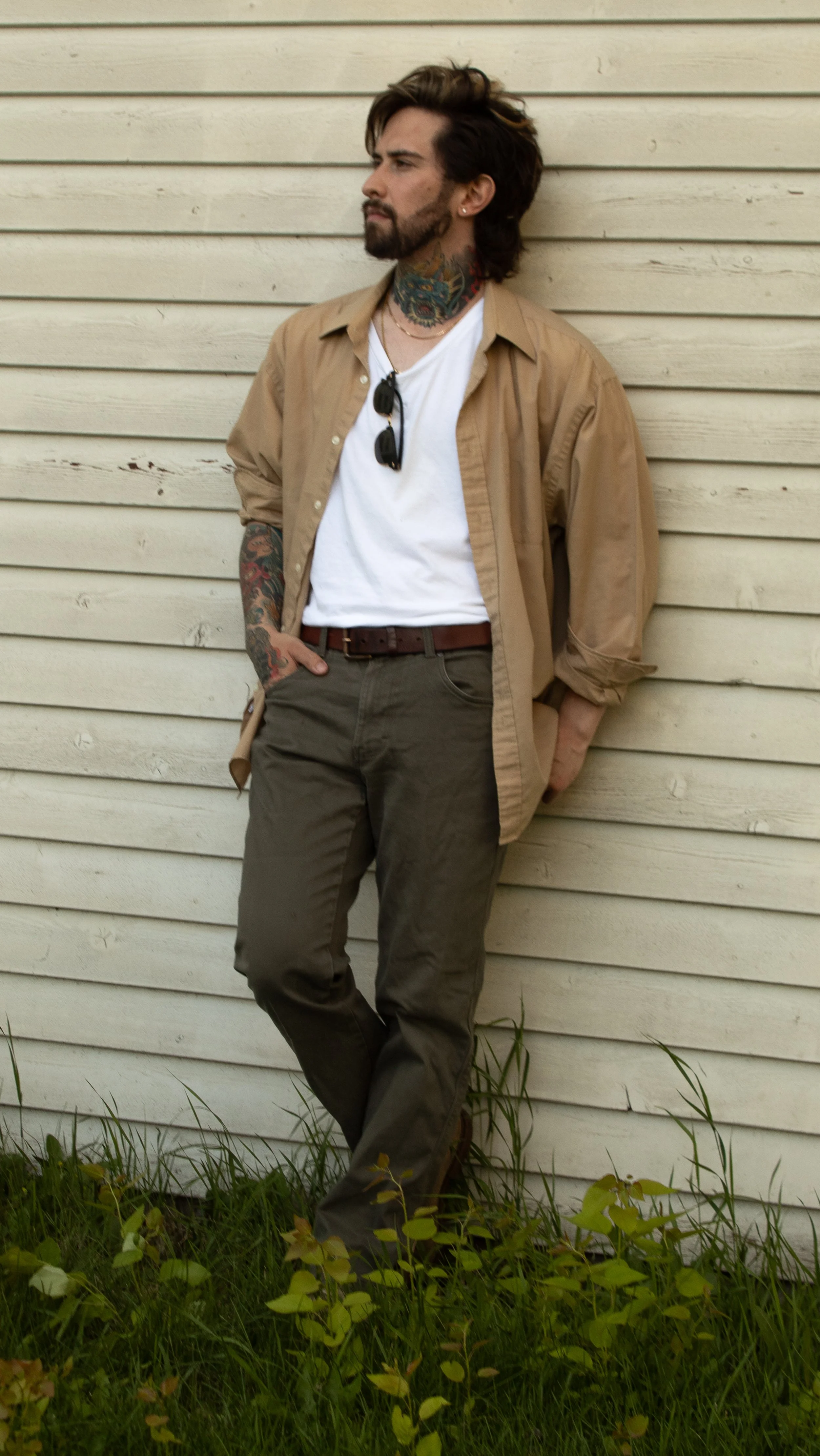 A man with tattoos and beard leaning against a wooden wall, wearing a tan shirt, white T-shirt, and dark pants.