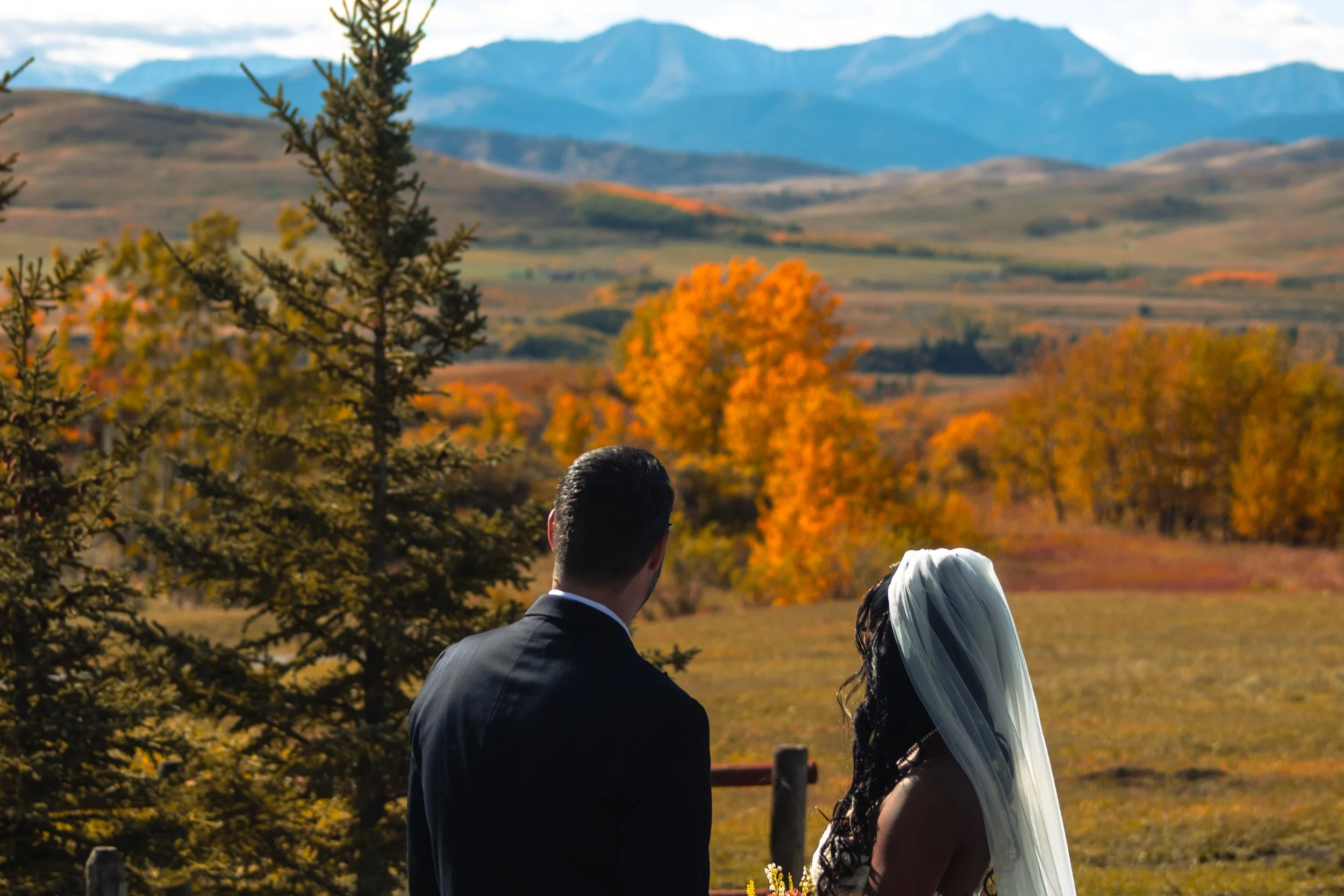 A bride and groom standing outdoors during autumn, with trees in fall colors and mountains in the background.