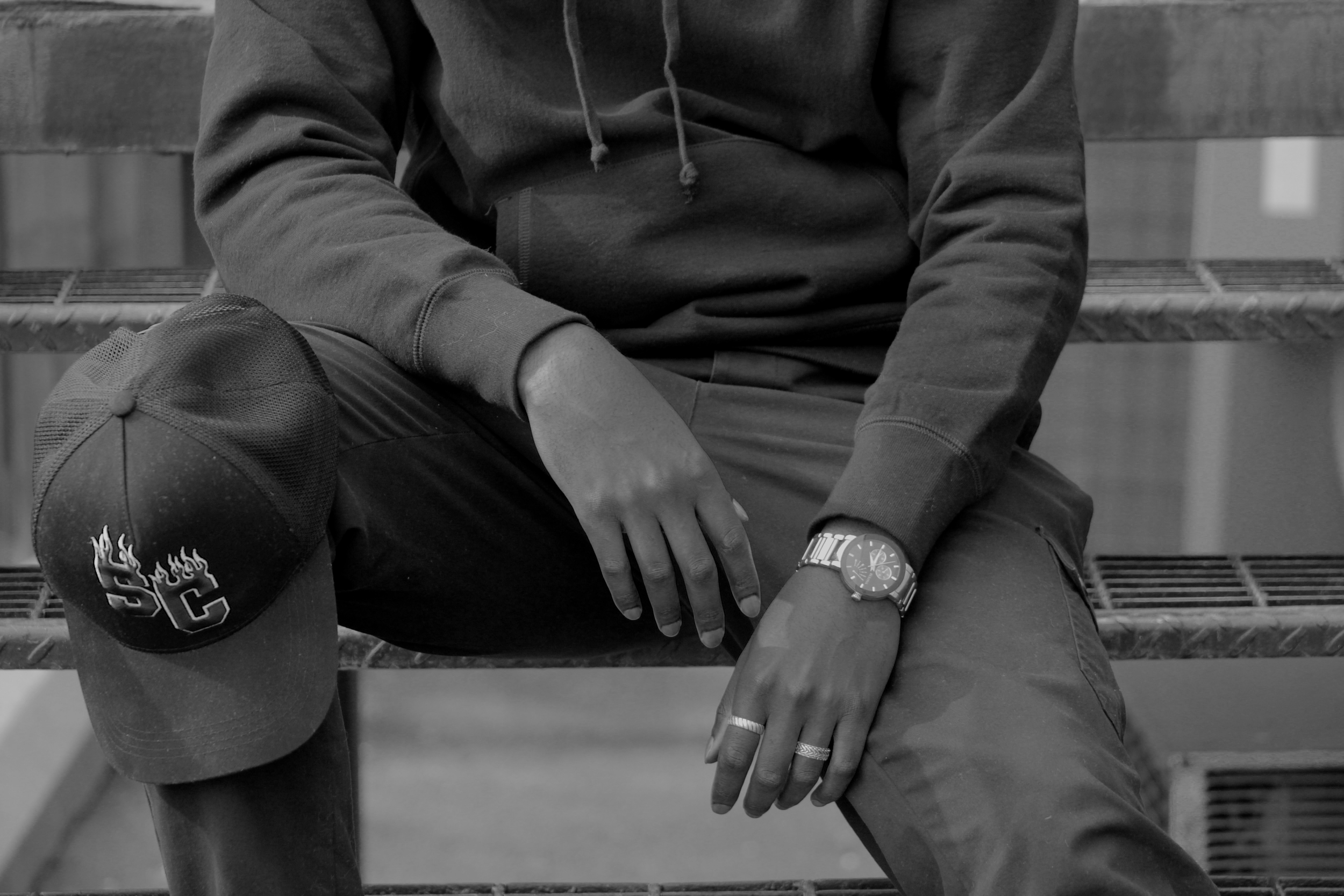 Person sitting on a bench wearing a hoodie, watch, rings, and a cap with 'SC' and flames logo, resting hands on their knees.