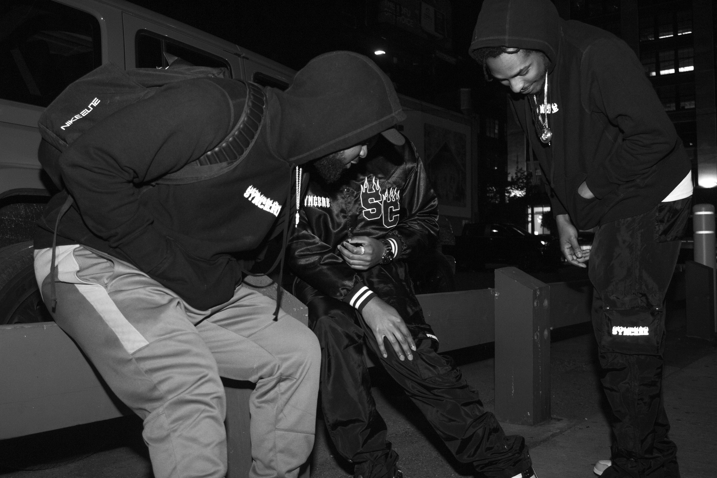 Three men sitting on a bench outdoors at night, engaging in conversation. All are dressed in hoodies and casual clothing.