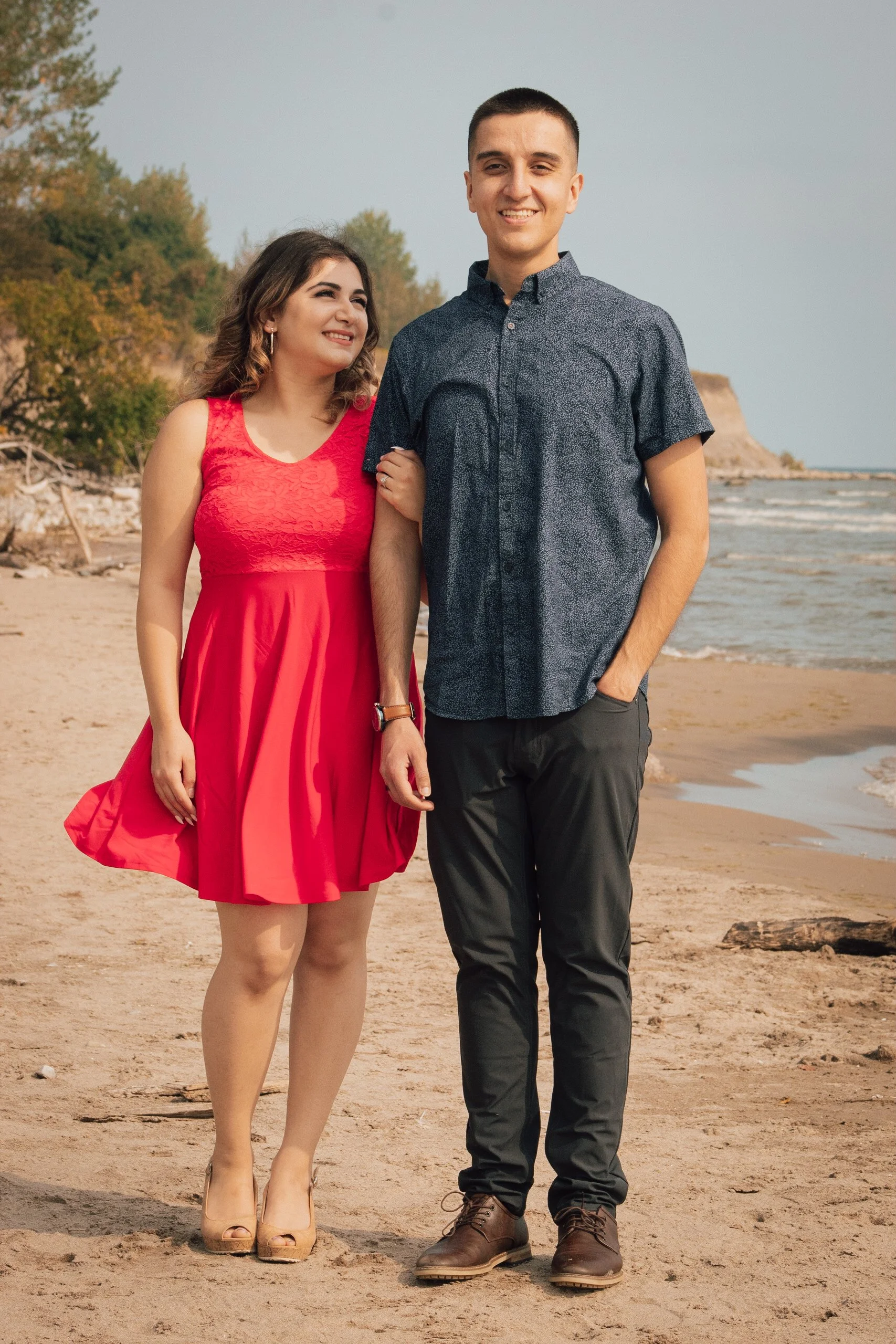 A smiling woman in a red dress and heels, and a man in a blue button-up shirt and dark pants, standing arm in arm on a sandy beach with trees and cliffs in the background.