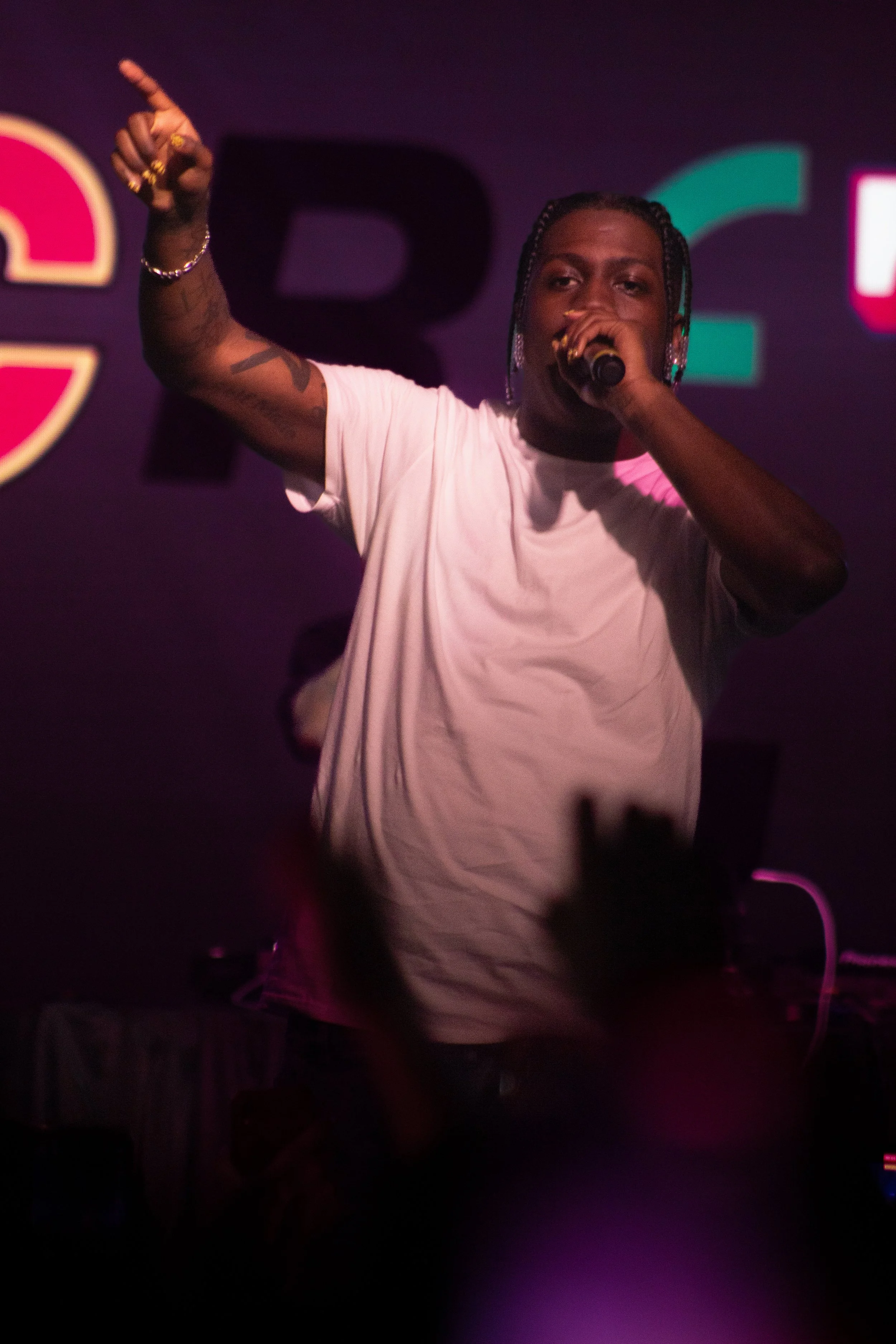Person with braids and jewelry holding a microphone, performing on stage with a dark background and colorful lights.