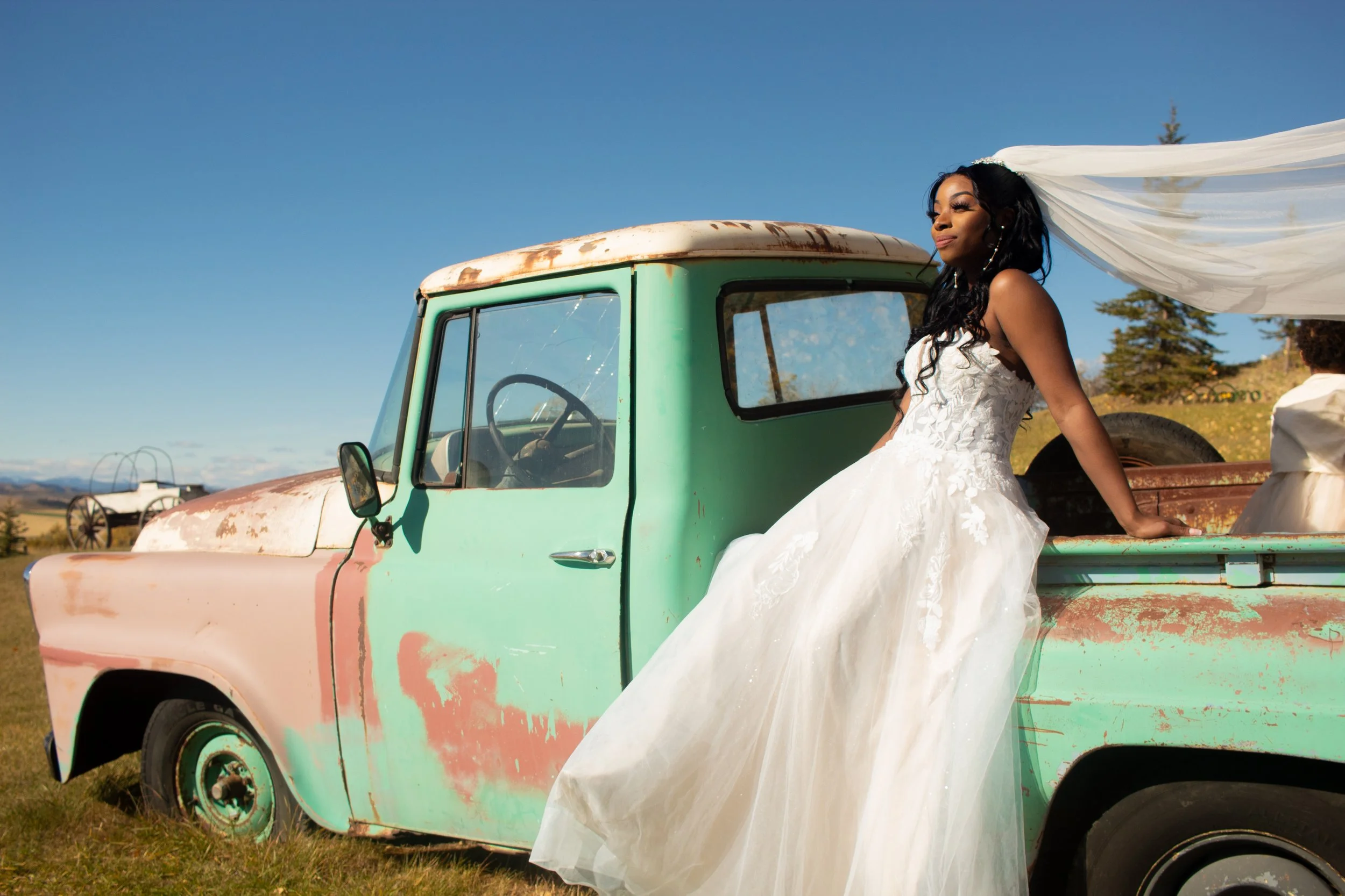A woman in a white wedding dress sitting on the bed of an old rusted turquoise pickup truck with a white veil flowing behind her, outdoors under a clear blue sky.
