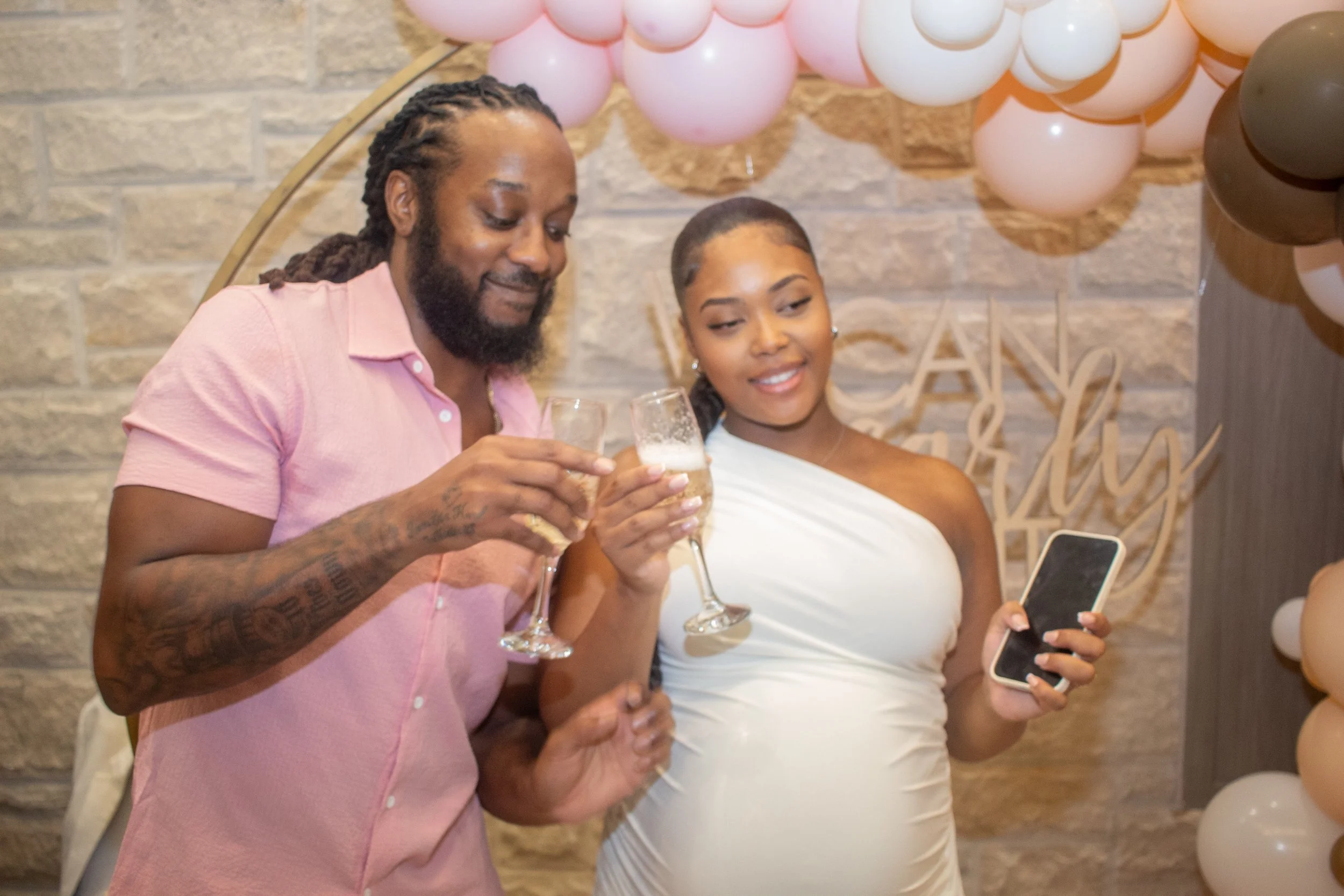 A man and a woman at a celebration, holding glasses of champagne and looking at a cellphone, with pink and gray balloons in the background.