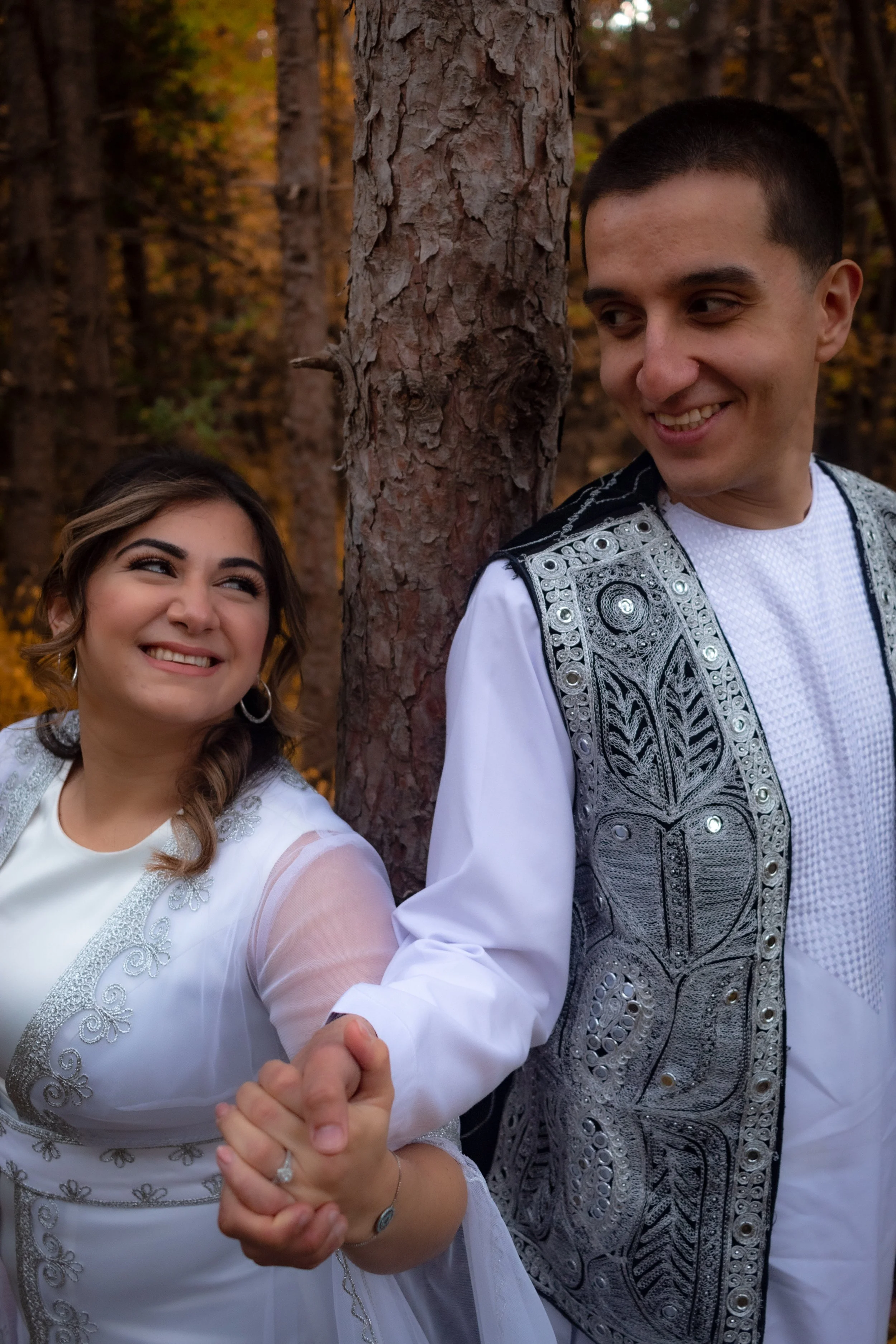 A smiling couple dressed in traditional attire, holding hands and standing in a forest with autumn leaves.