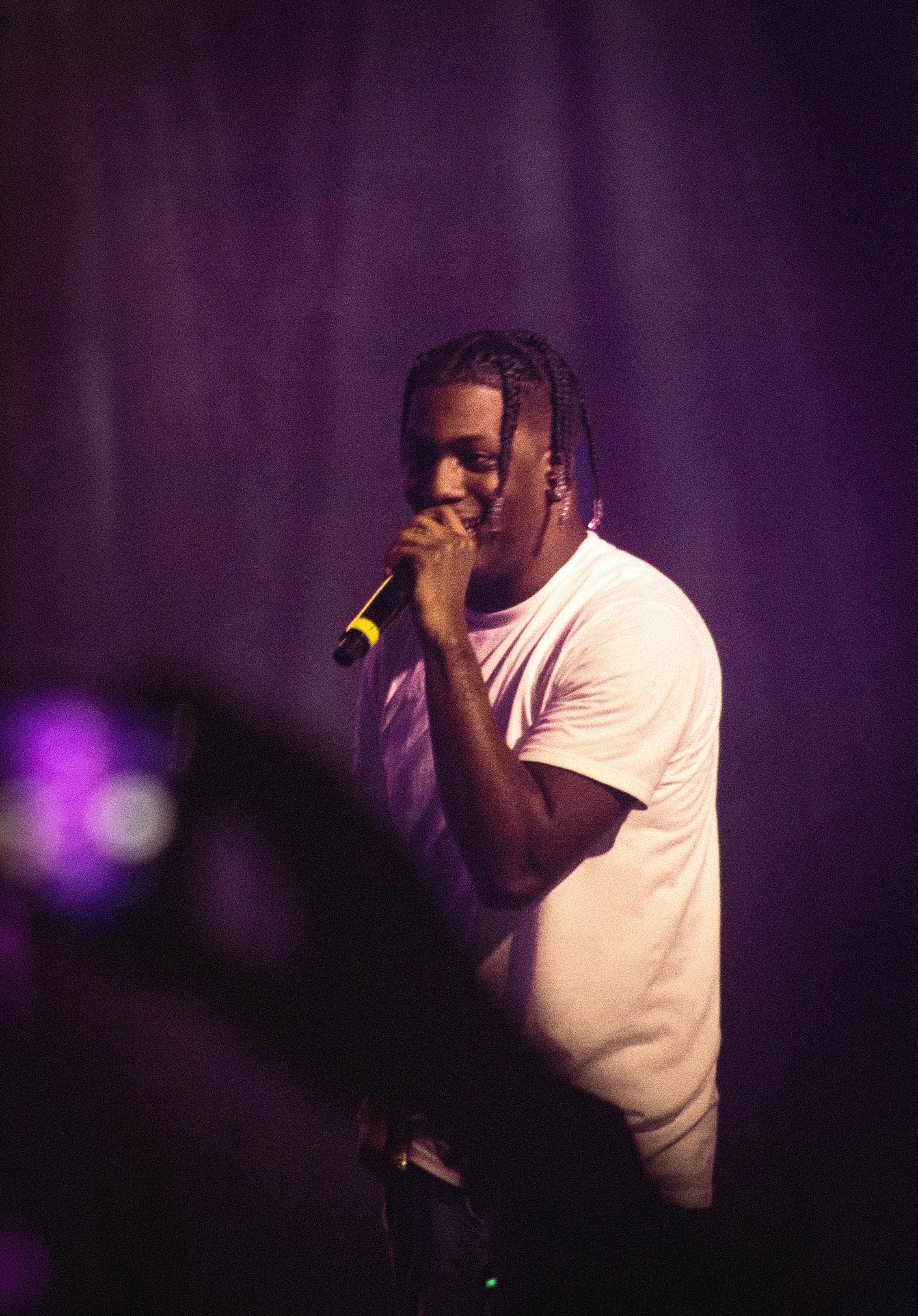 A young man with braided hair singing into a microphone on stage, wearing a white t-shirt, with purple stage lighting in the background.