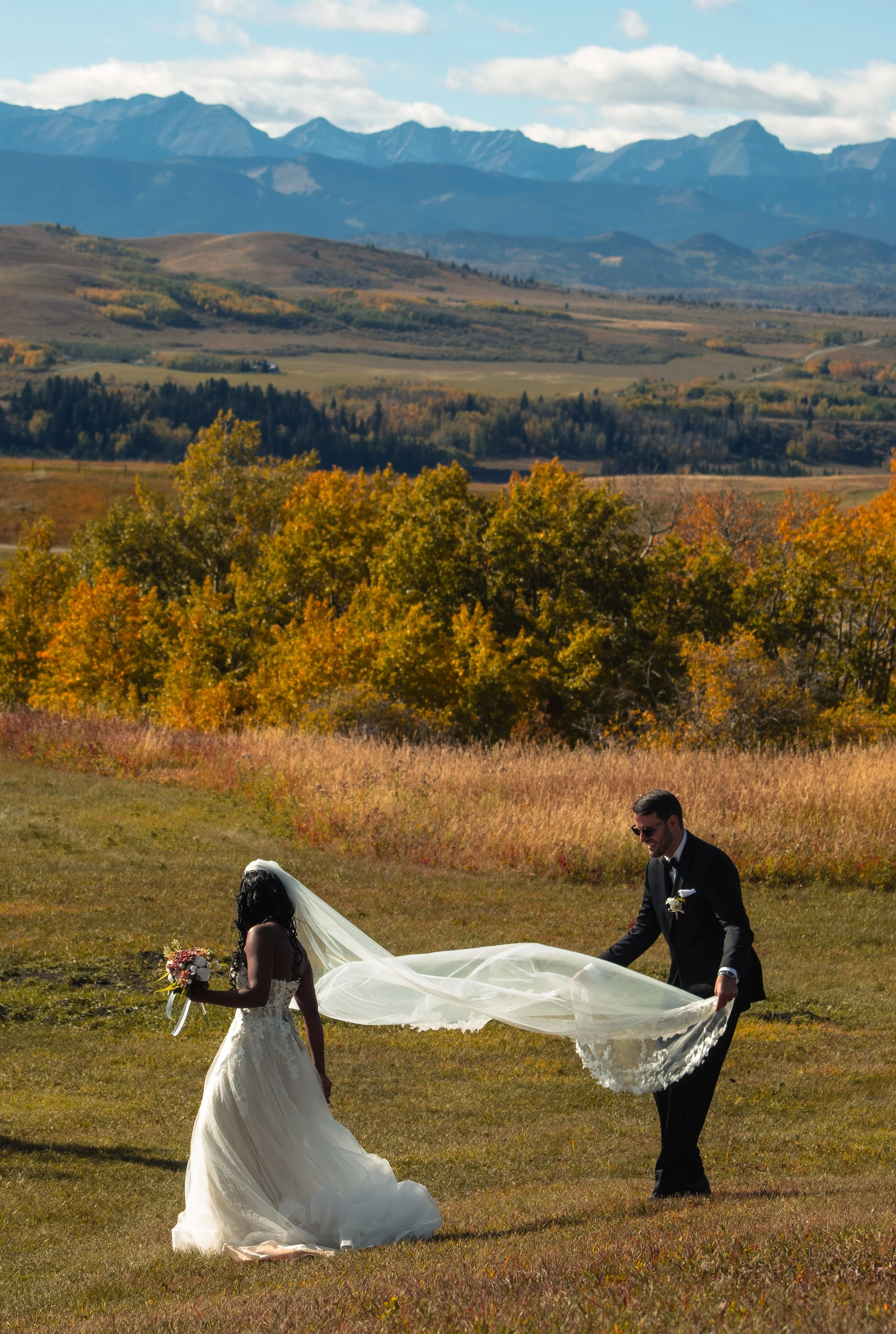 A bride and groom holding hands and smiling outdoors during a wedding ceremony, with a scenic mountain range and colorful autumn trees in the background.