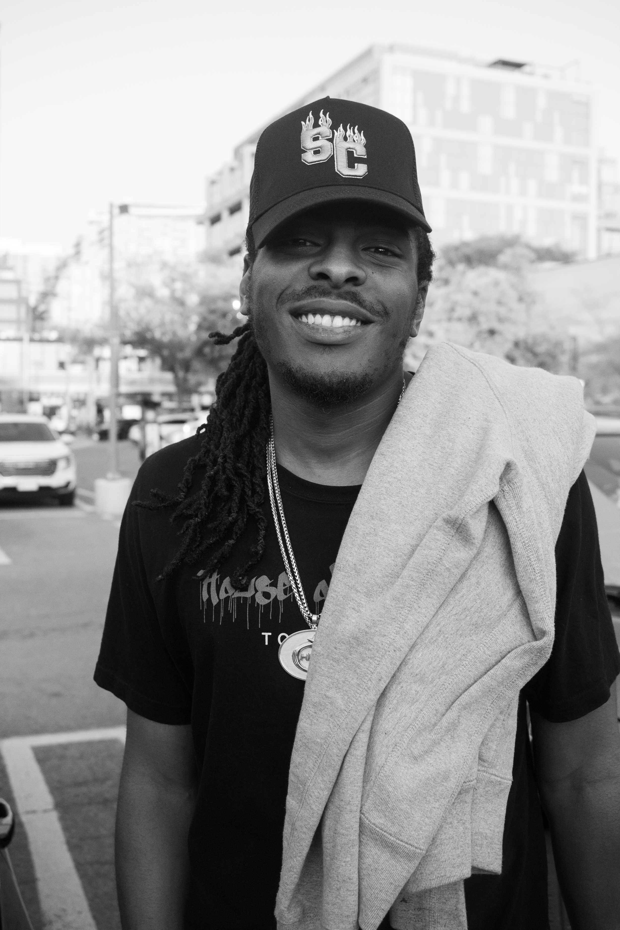 A smiling man with dreadlocks wearing a baseball cap with flames and letters on it. He has a towel or sweatshirt draped over his shoulder and is standing outdoors in a parking lot with modern buildings in the background.
