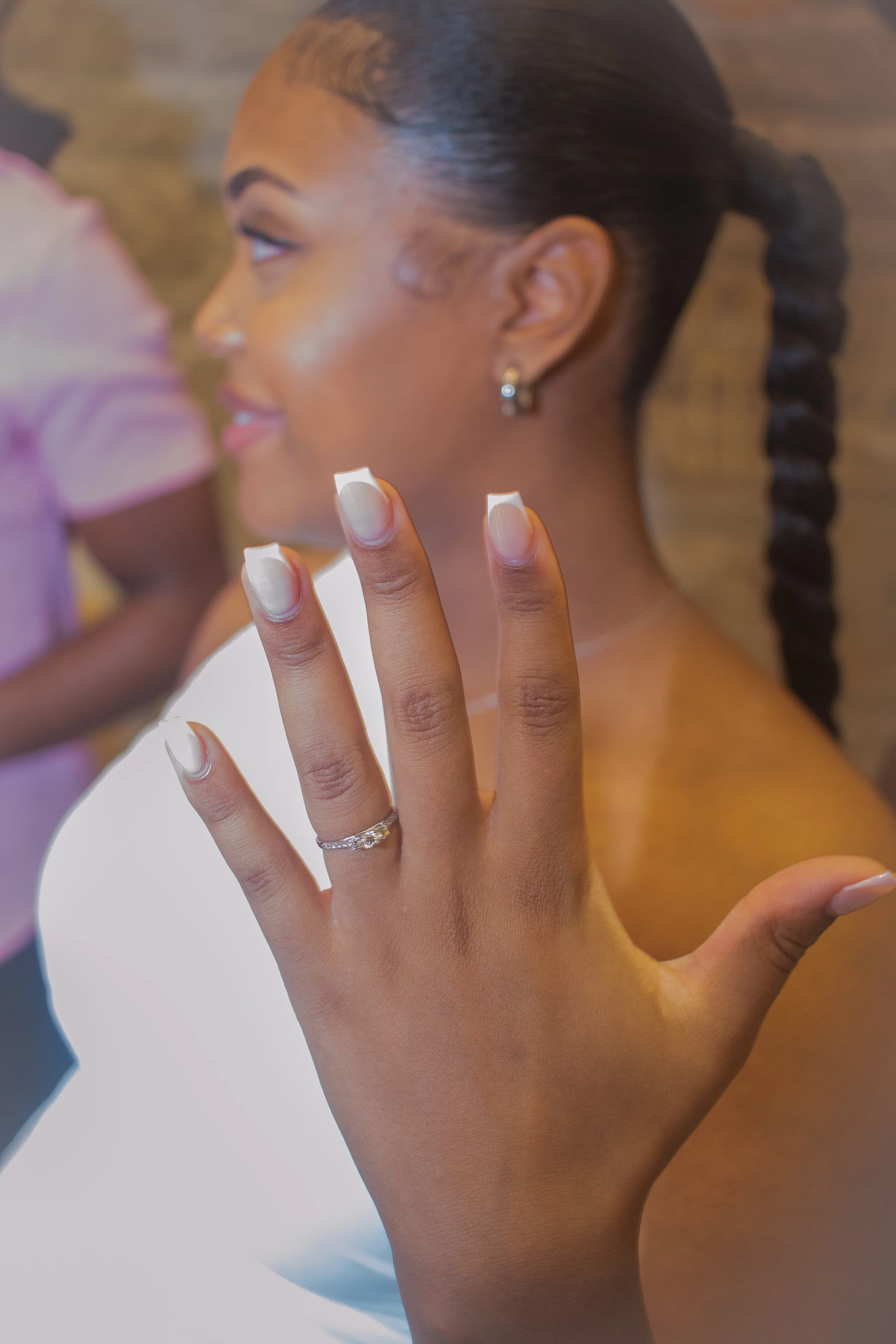 Close-up of a woman with a braid hairstyle, showing her face in profile, with a hand in the foreground displaying a ring and well-manicured nails.