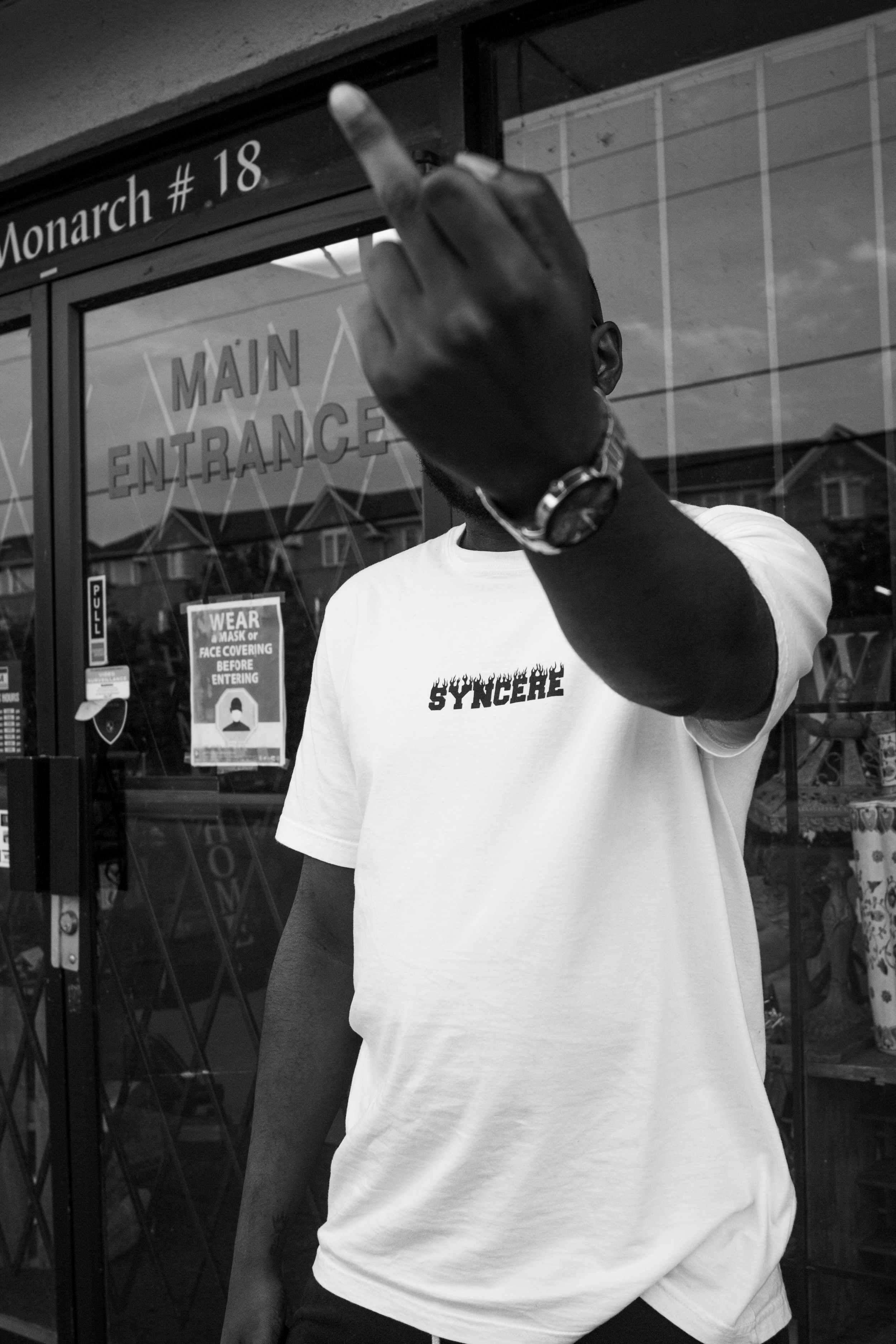 A person in a white t-shirt with the word 'SYNCHRE' printed on it stands outside a building with glass doors. The person is holding up their middle finger, obscuring part of their face. The building has signs indicating it is the main entrance, and t