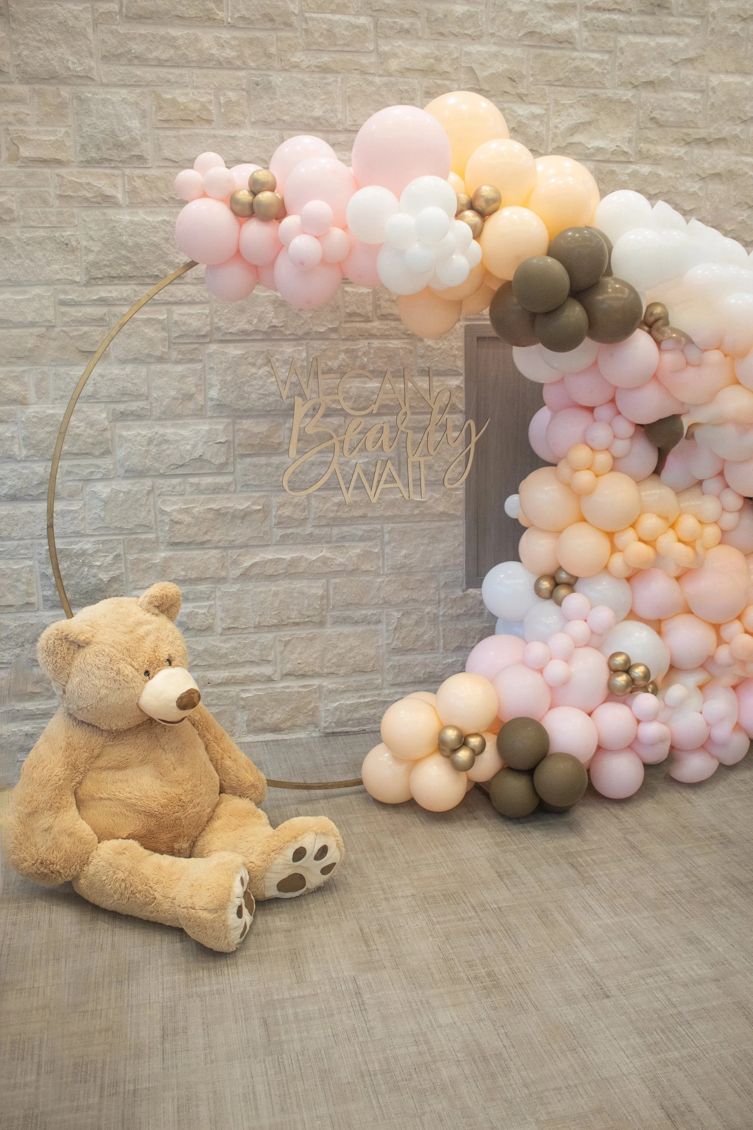 A large teddy bear sitting on a grey textured carpet in front of a balloon arch in pastel pink, white, yellow, gold, and dark green, with a wooden sign that reads 'We Can Bearly WAI' attached to the arch. The background is a beige brick wall.