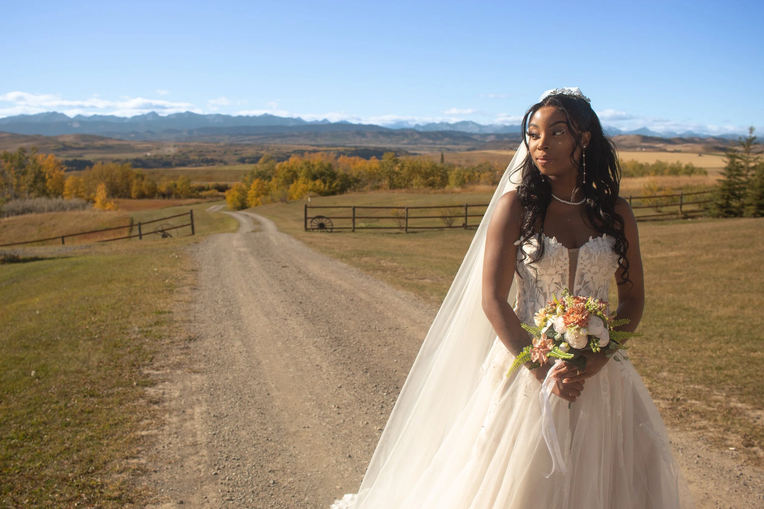 A woman in a white wedding dress holding a bouquet stands outdoors on a dirt road, with a scenic landscape of rolling hills, trees with autumn foliage, a wooden fence, and mountains in the background under a partly cloudy sky.