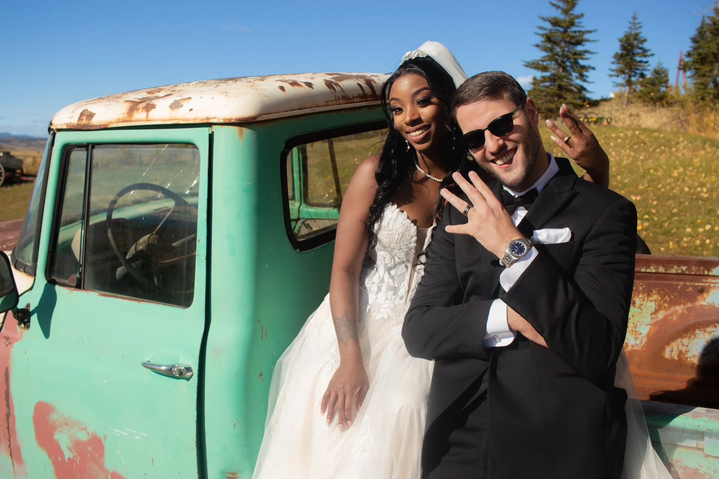A newlywed couple takes a selfie in front of a vintage rusted green truck outdoors during daytime, with a backdrop of trees and a clear sky.
