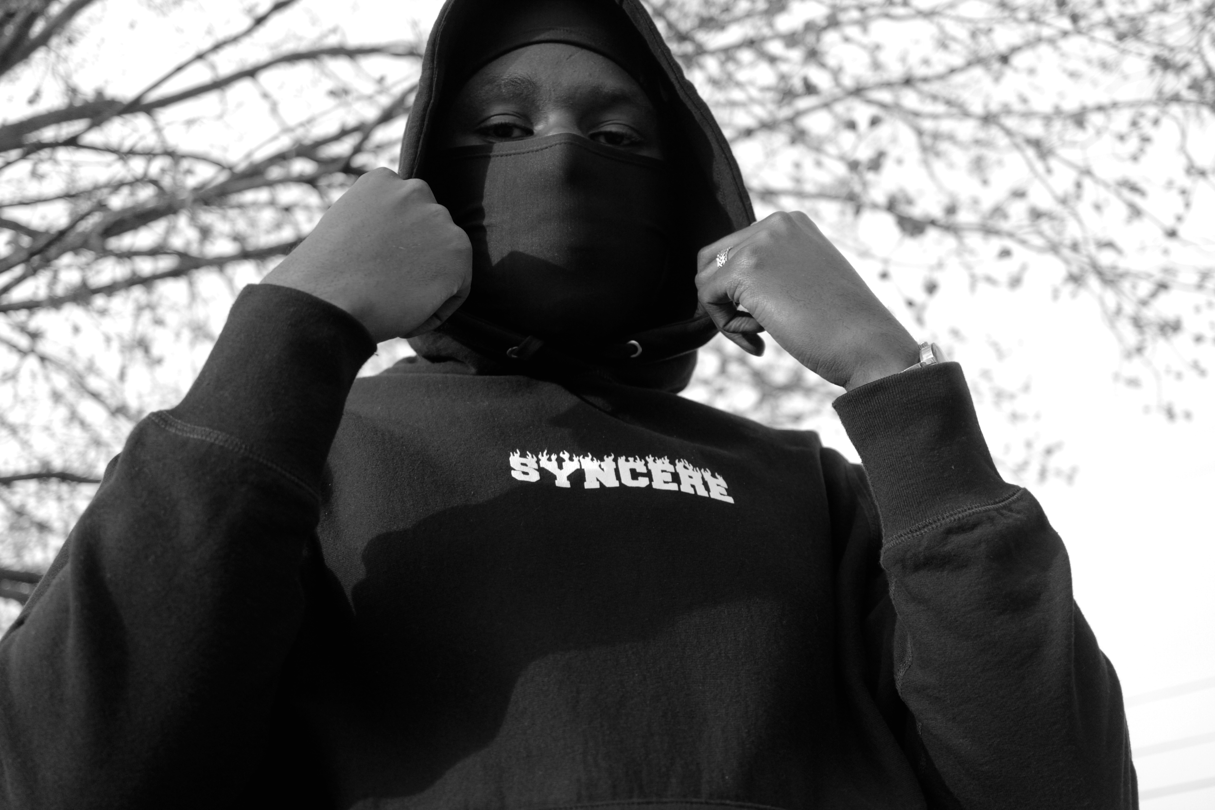 A person wearing a black hoodie, black face covering, and rings on their fingers, adjusting their hoodie outdoors under leafless trees.