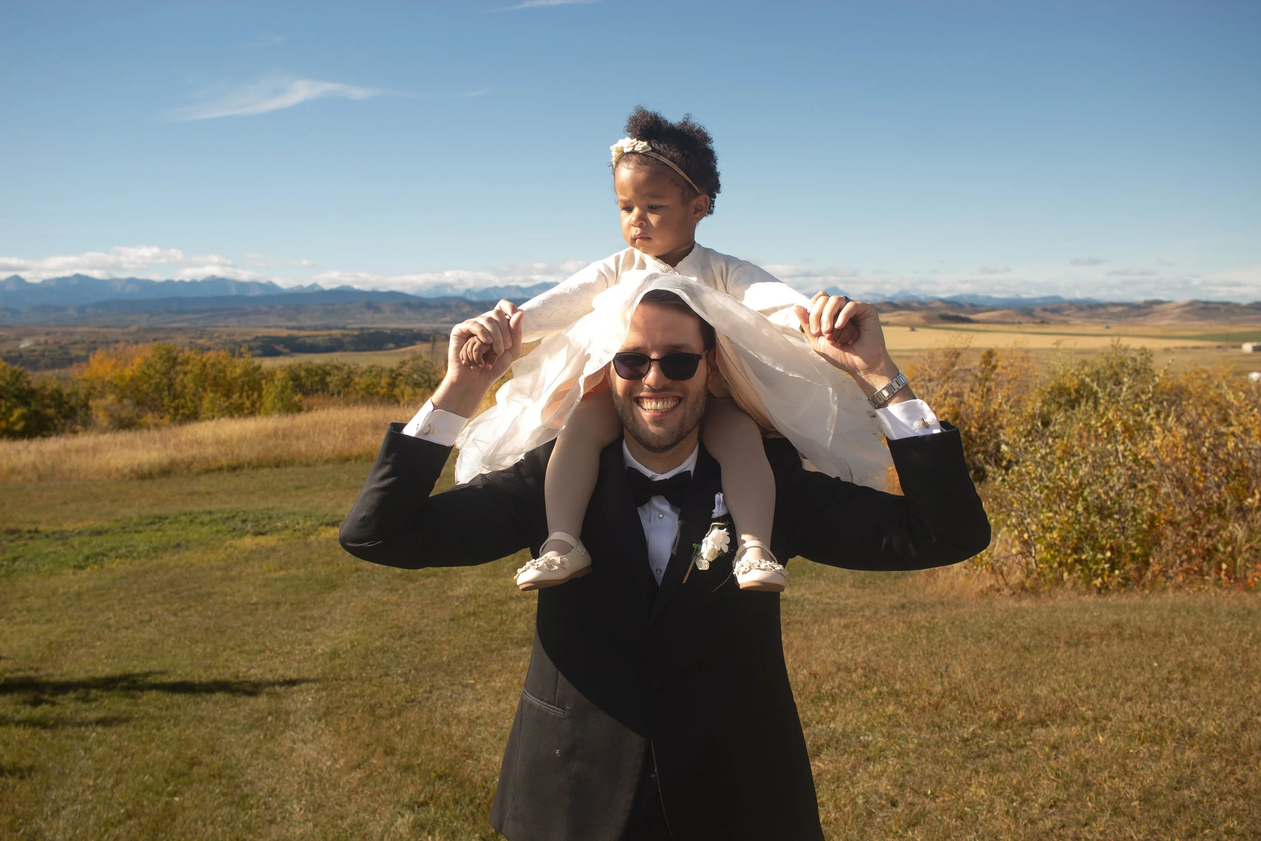 A man in a black tuxedo carrying a young girl on his shoulders outdoors with mountains and a blue sky in the background.