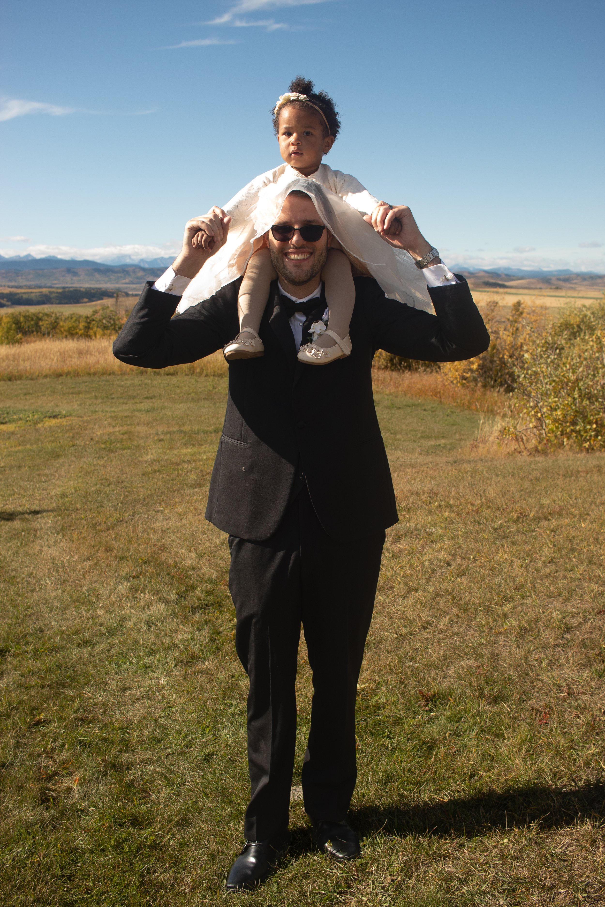 A man wearing a black suit and sunglasses stands outdoors on grass, carrying a young girl on his shoulders, with mountains and a blue sky in the background.