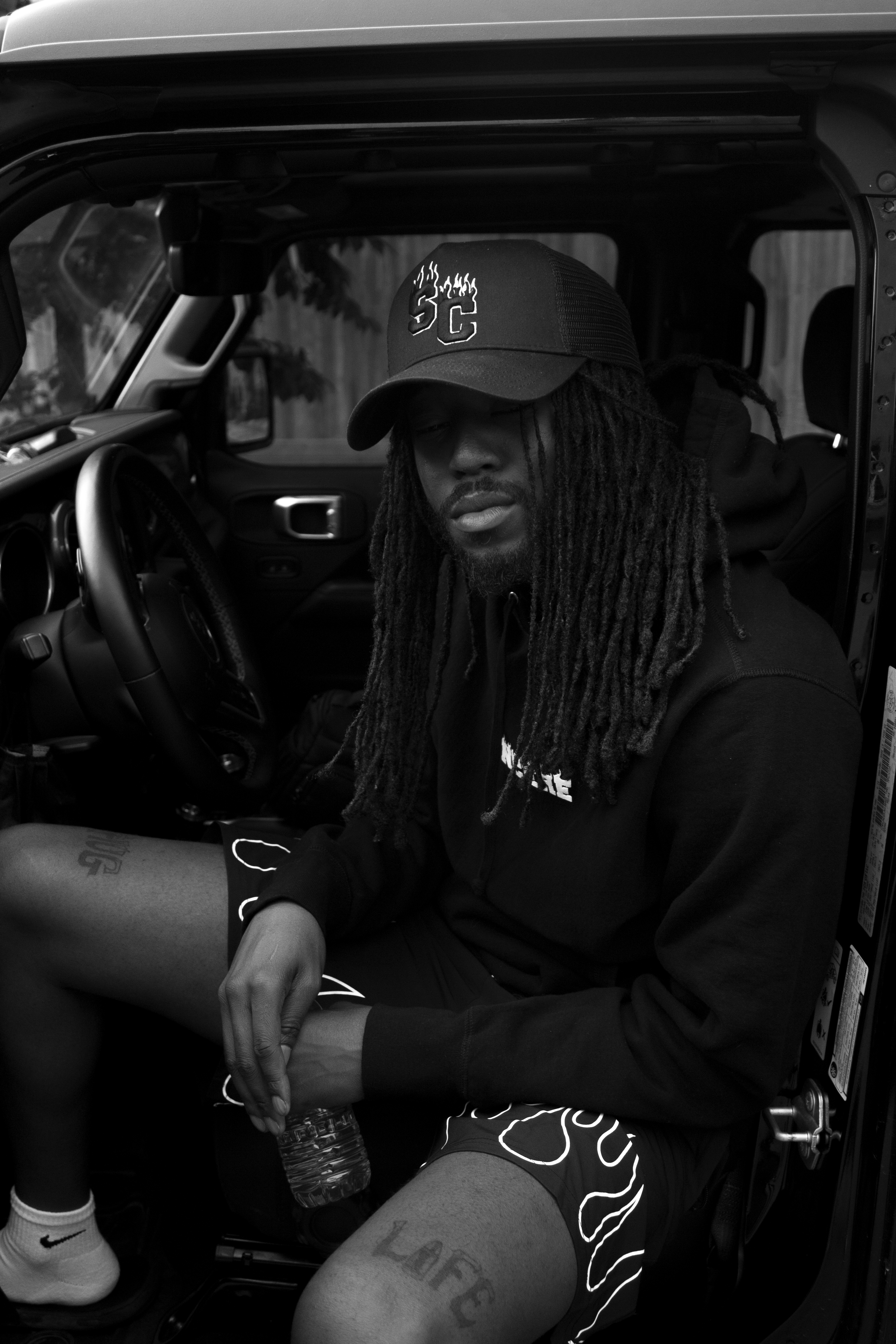 Young man with long dreadlocks wearing a baseball cap, black hoodie, and shorts, sitting inside a vehicle, holding a water bottle.