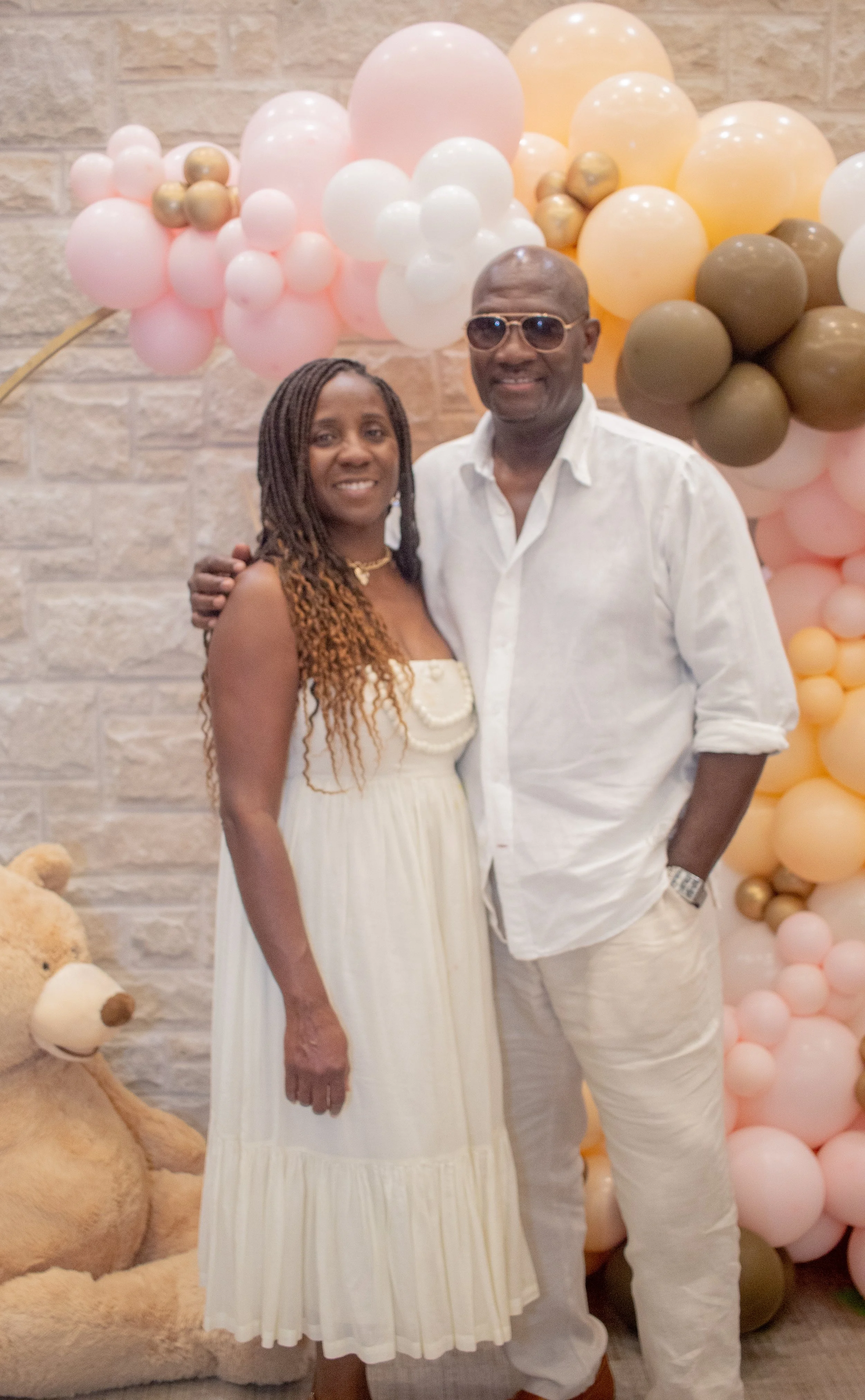 A couple standing in front of balloon decorations with pastel colors, including pink, white, beige, and gray, against a brick wall background. The woman is wearing a white dress with pearl details, and the man is dressed in a white shirt and white pa