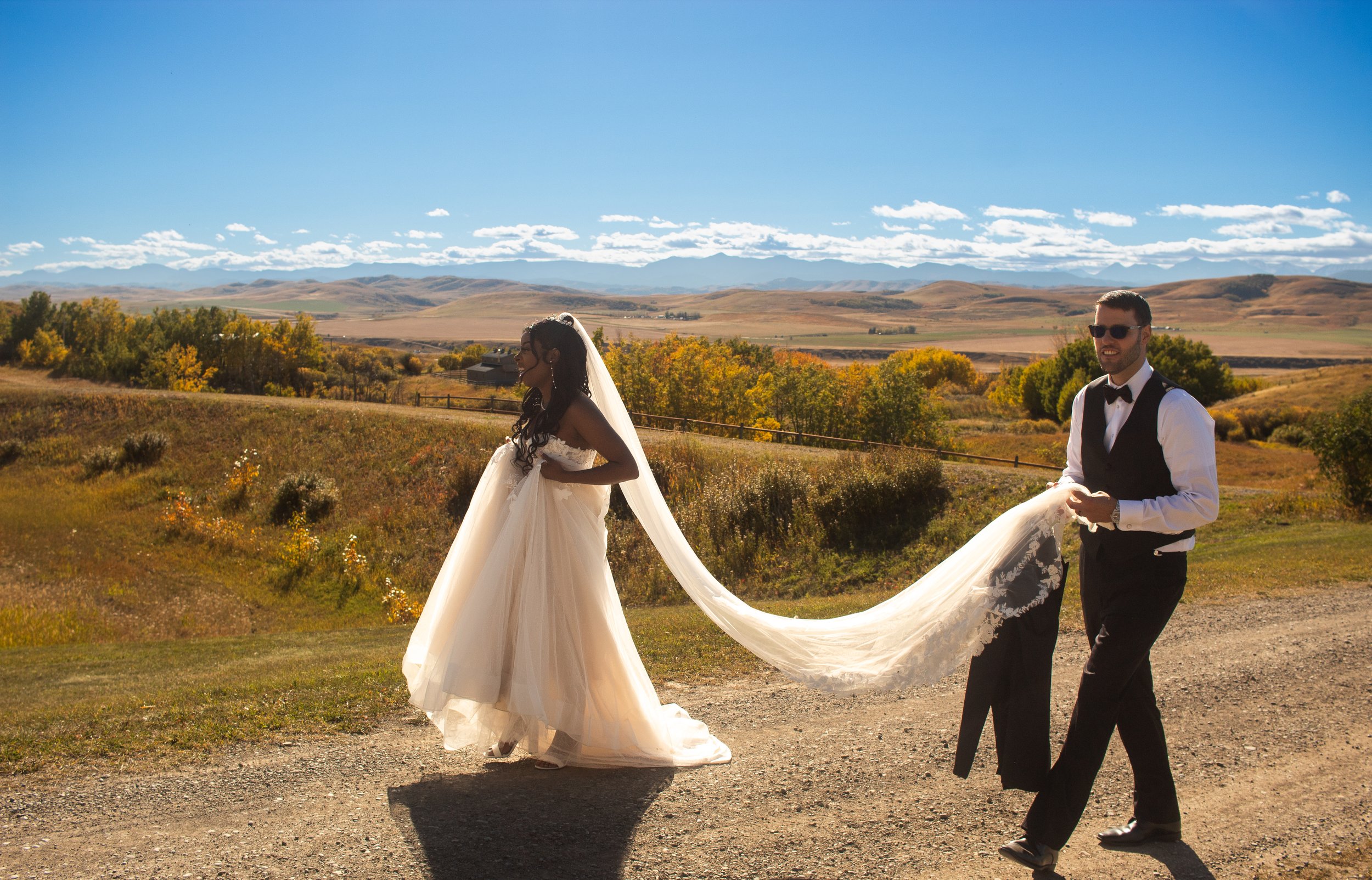 A bride and groom walking outdoors on a sunny day with a landscape of rolling hills, trees with fall foliage, and a mountain range in the distance.