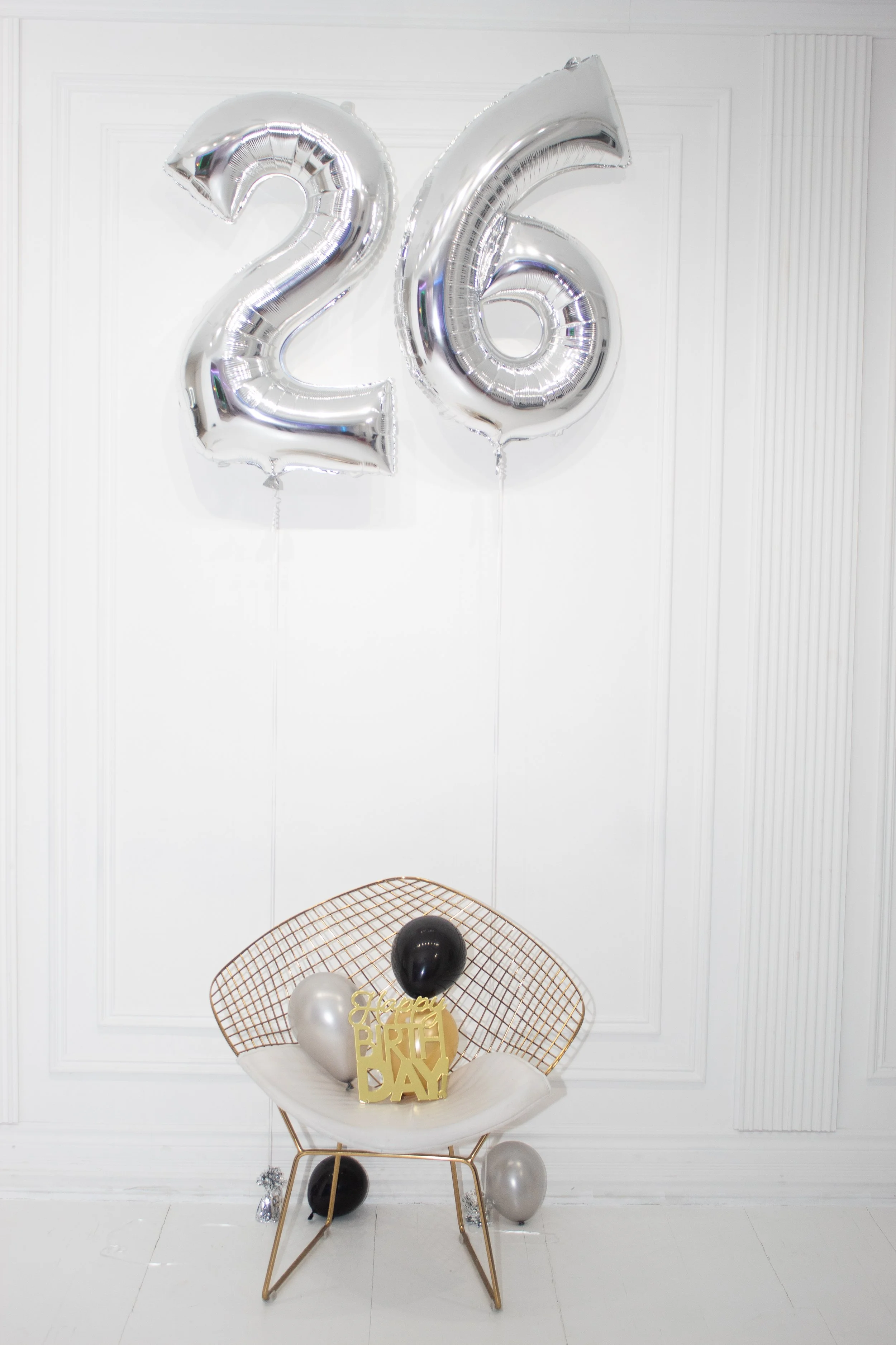 Silver number balloons displaying '26' hung on a white wall, with a decorative seating area beneath that includes black, silver, and white balloons, a gold 'Happy Birthday' sign, and a gold wire mesh backdrop.