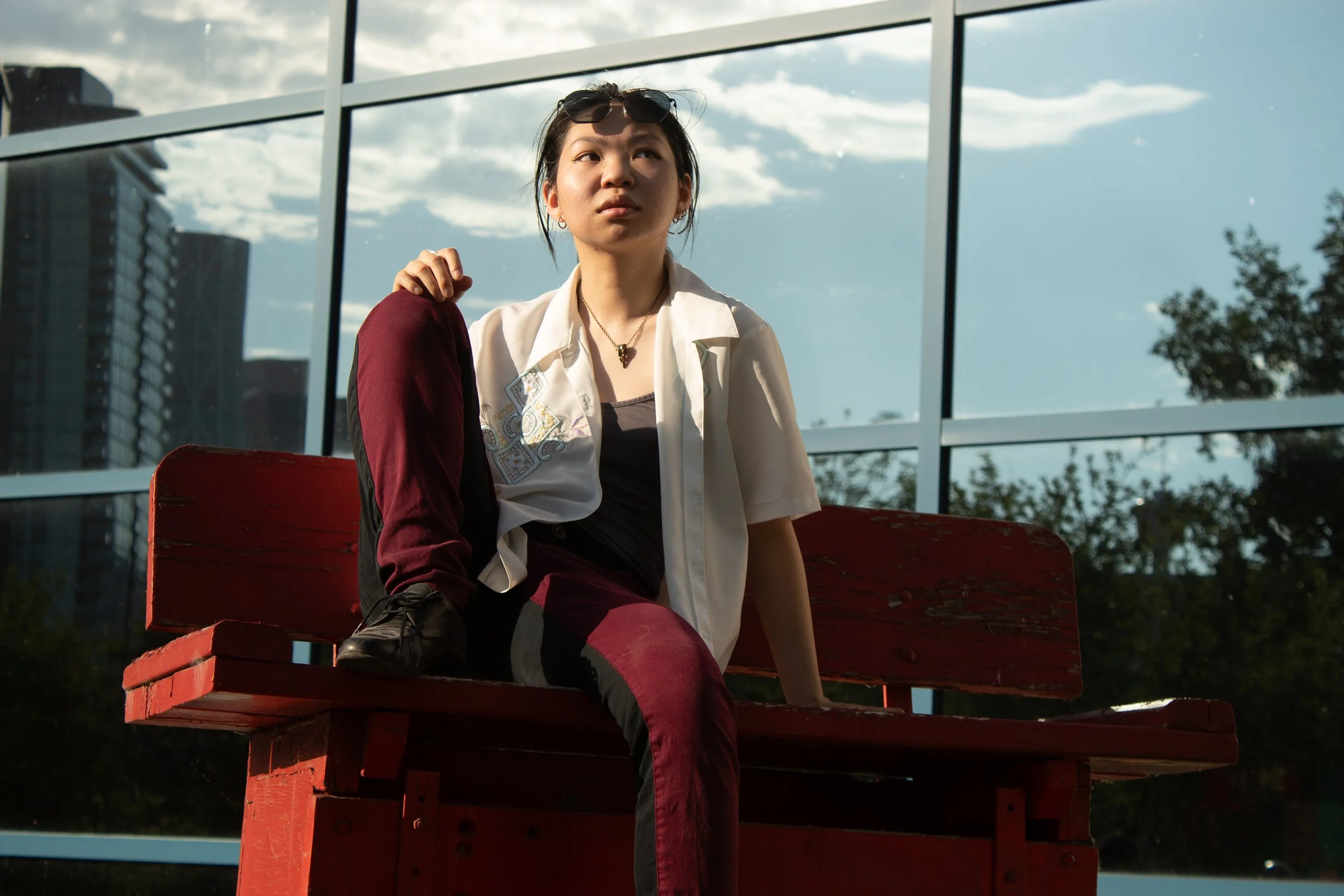A young woman with dark hair, sunglasses on her head, wearing a white shirt, black tank top, and maroon pants, sitting on a red bench outside with a modern glass building and cloudy sky in the background.