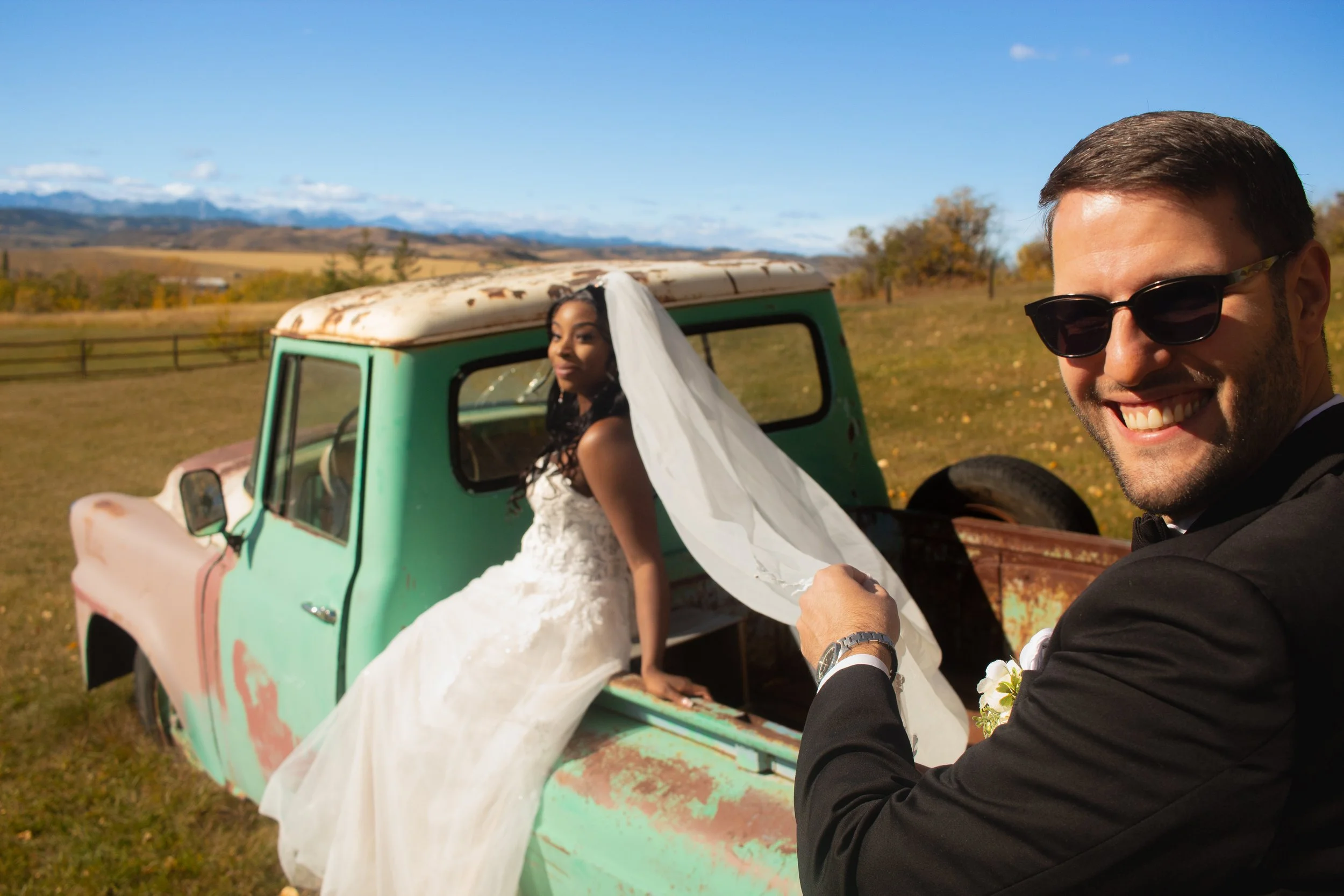 A man in sunglasses smiling as he takes a selfie with a woman in a wedding dress who is seated in an old, rusted green pickup truck in an open field during daytime with mountains in the background.