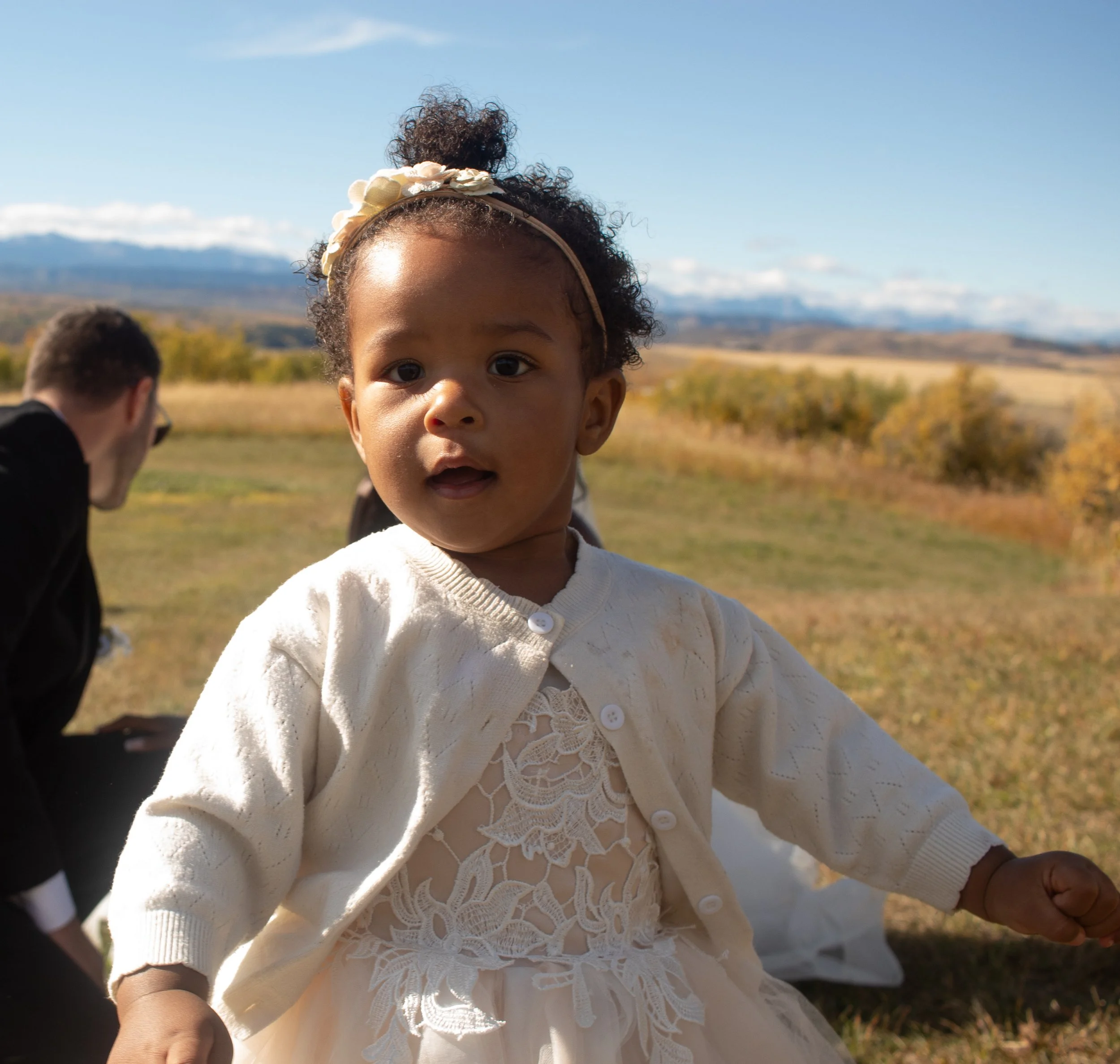 Close-up of a young girl with curly hair wearing a cream-colored, lace cardigan and a headband with fabric flowers, outdoors with open sky and distant mountains.