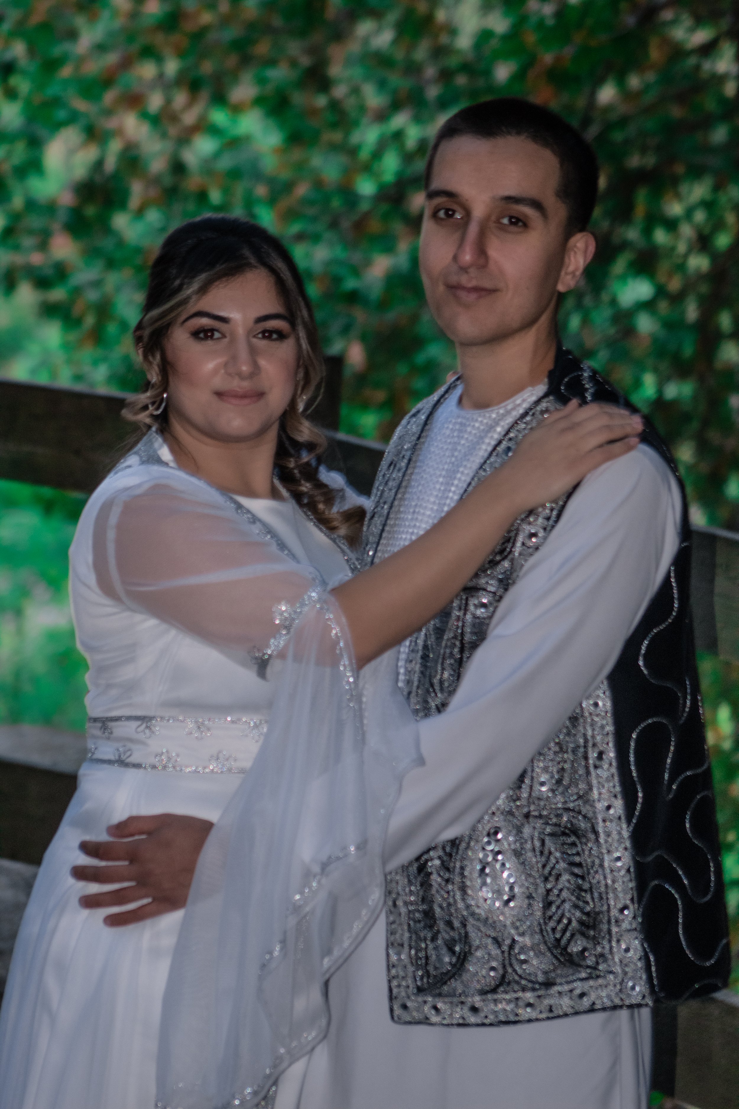 A couple embracing, dressed in elegant outfits, standing outdoors with green foliage in the background.
