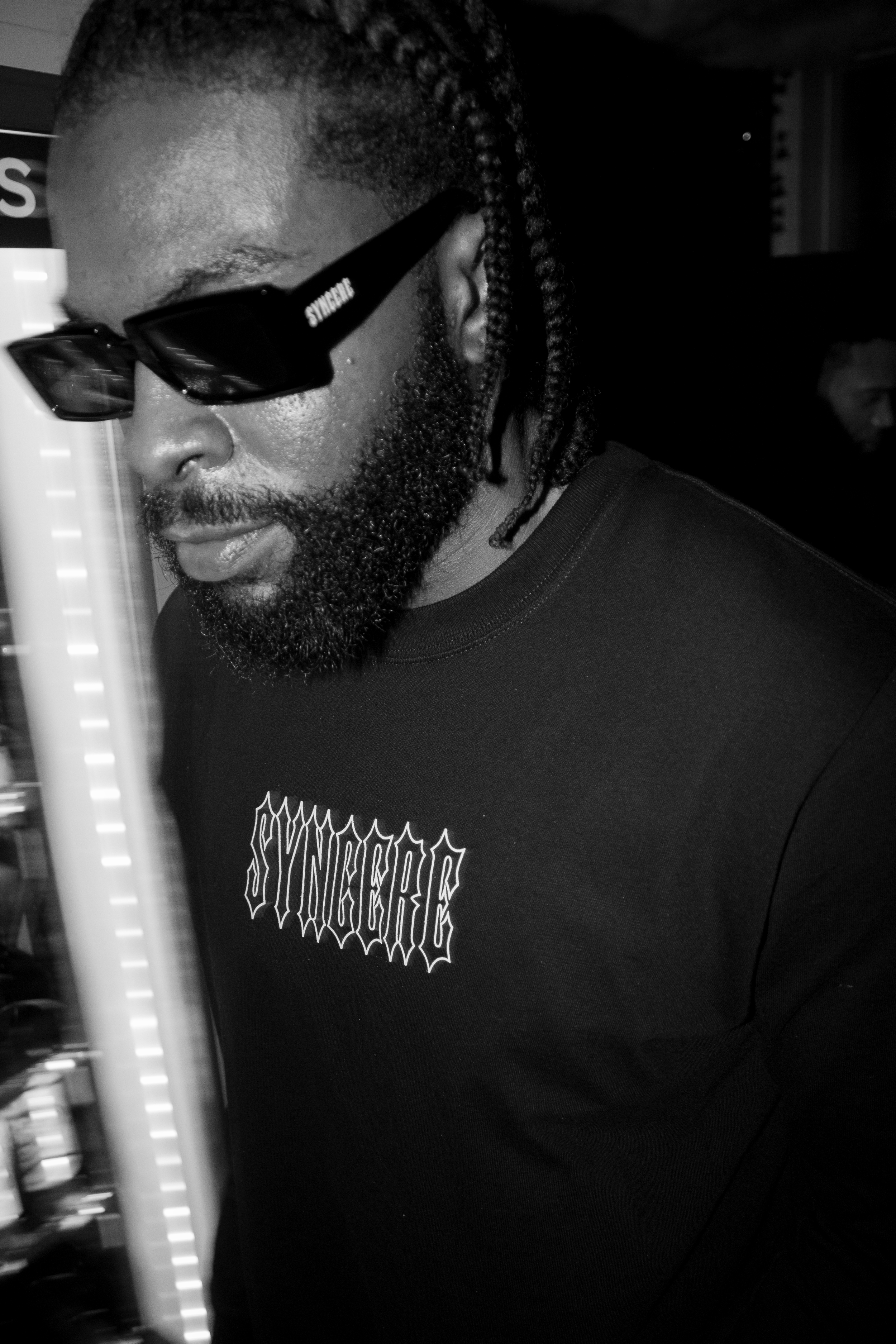 A man with a beard and glasses is standing indoors, wearing a black shirt with white text, and looking slightly downward.