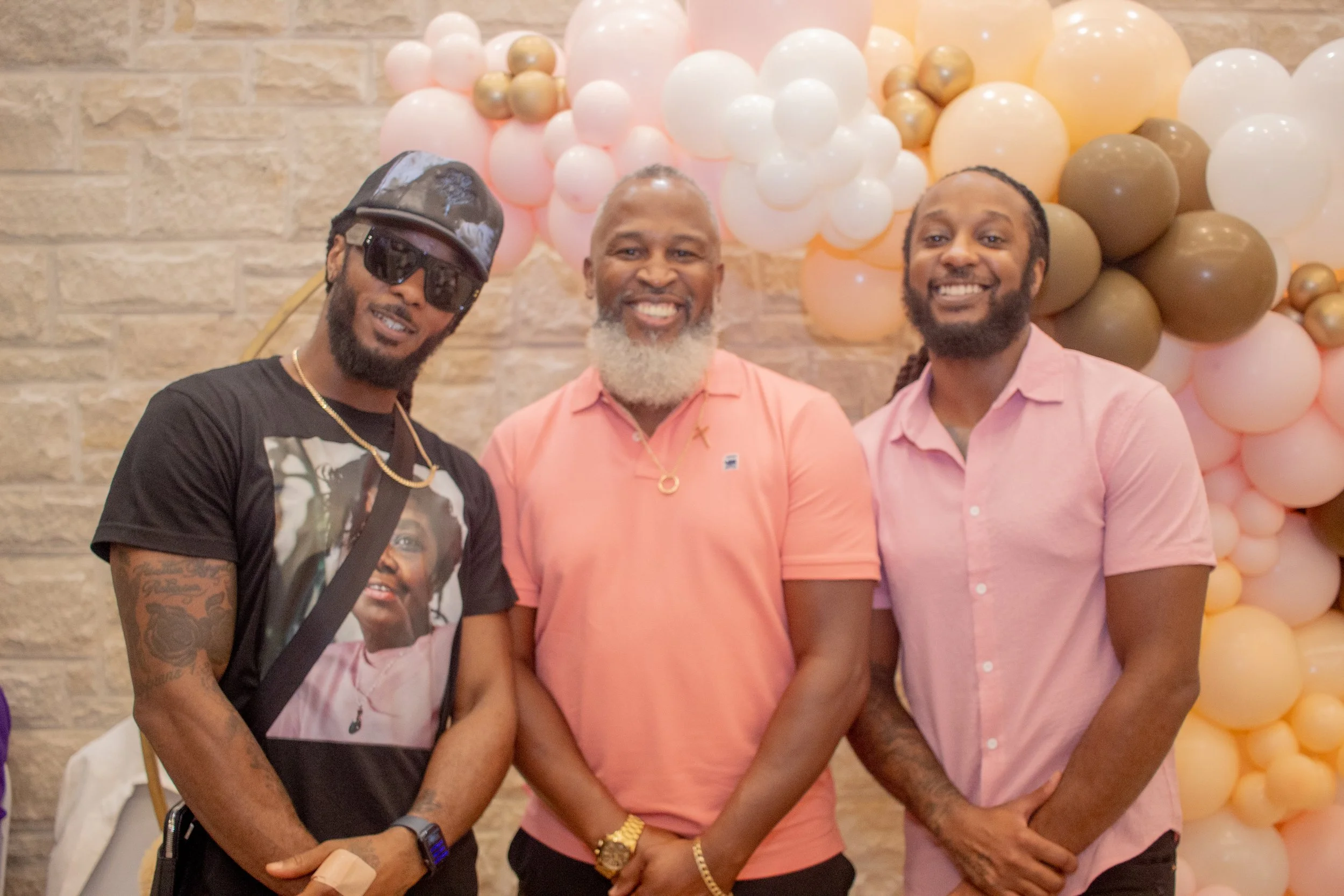 Three men smiling at a party in front of balloon decorations in pastel colors.